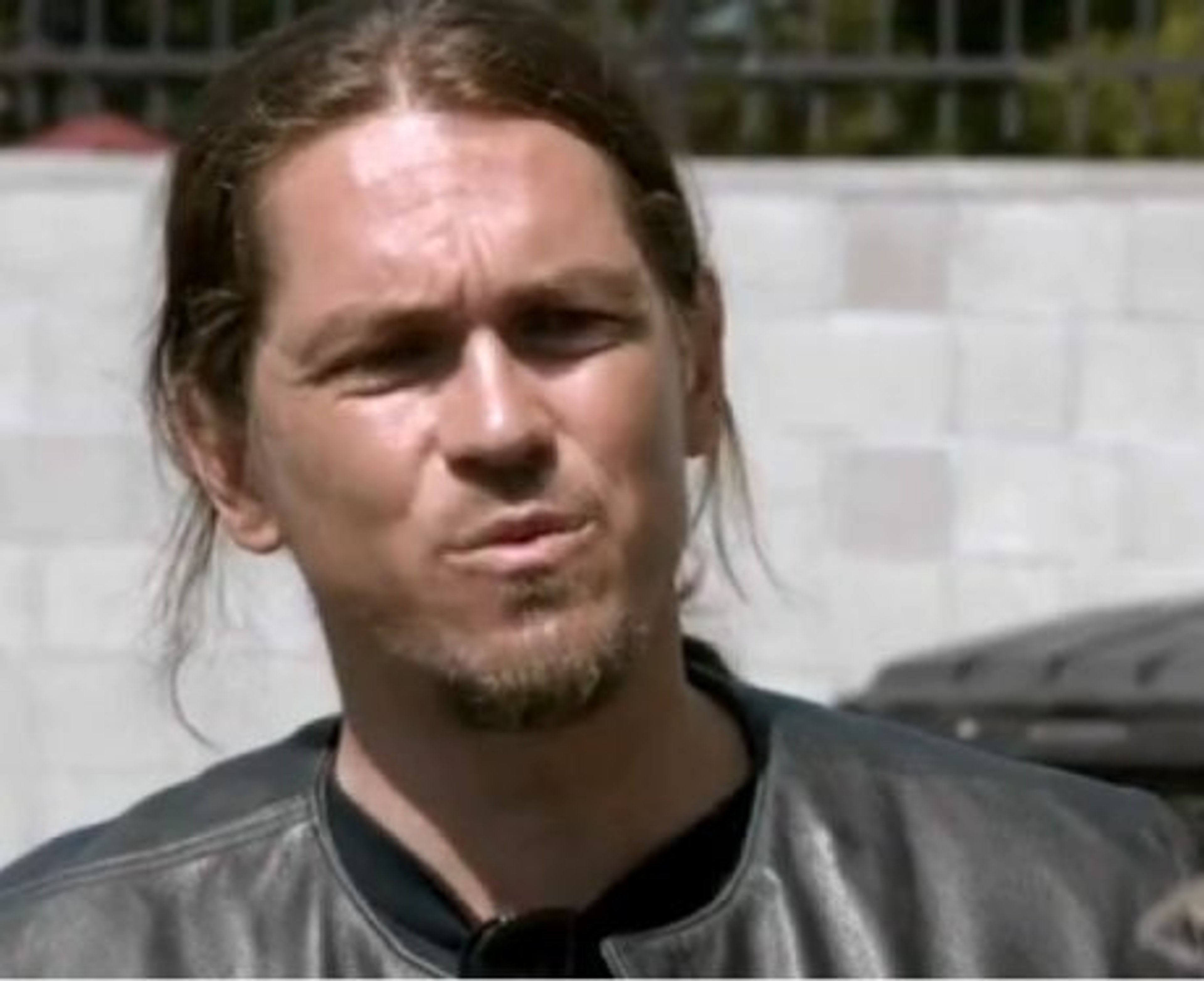 A middle-aged man with long hair and a beard, wearing a black leather jacket and having a tough, serious expression.
