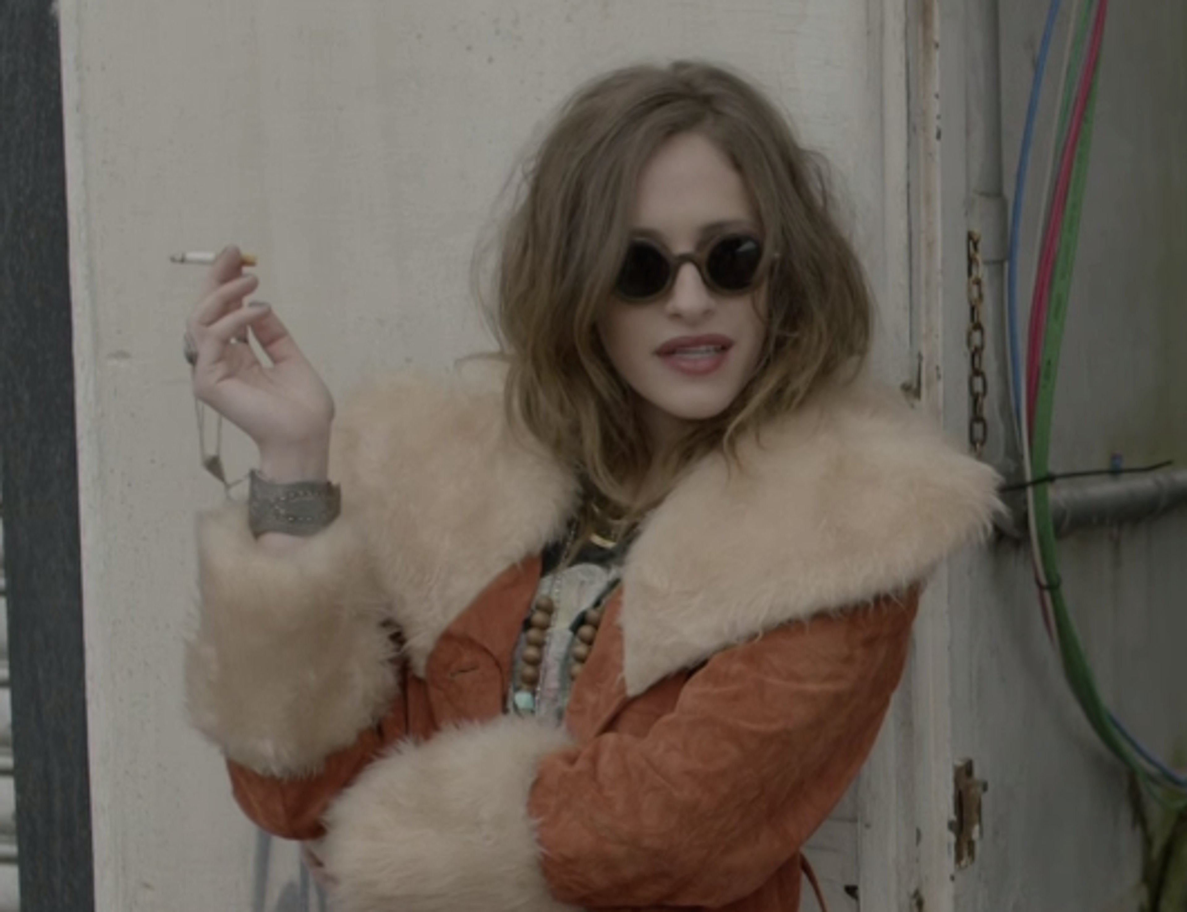A woman with long brown hair wearing an orange fur-trimmed jacket and holding a cigarette