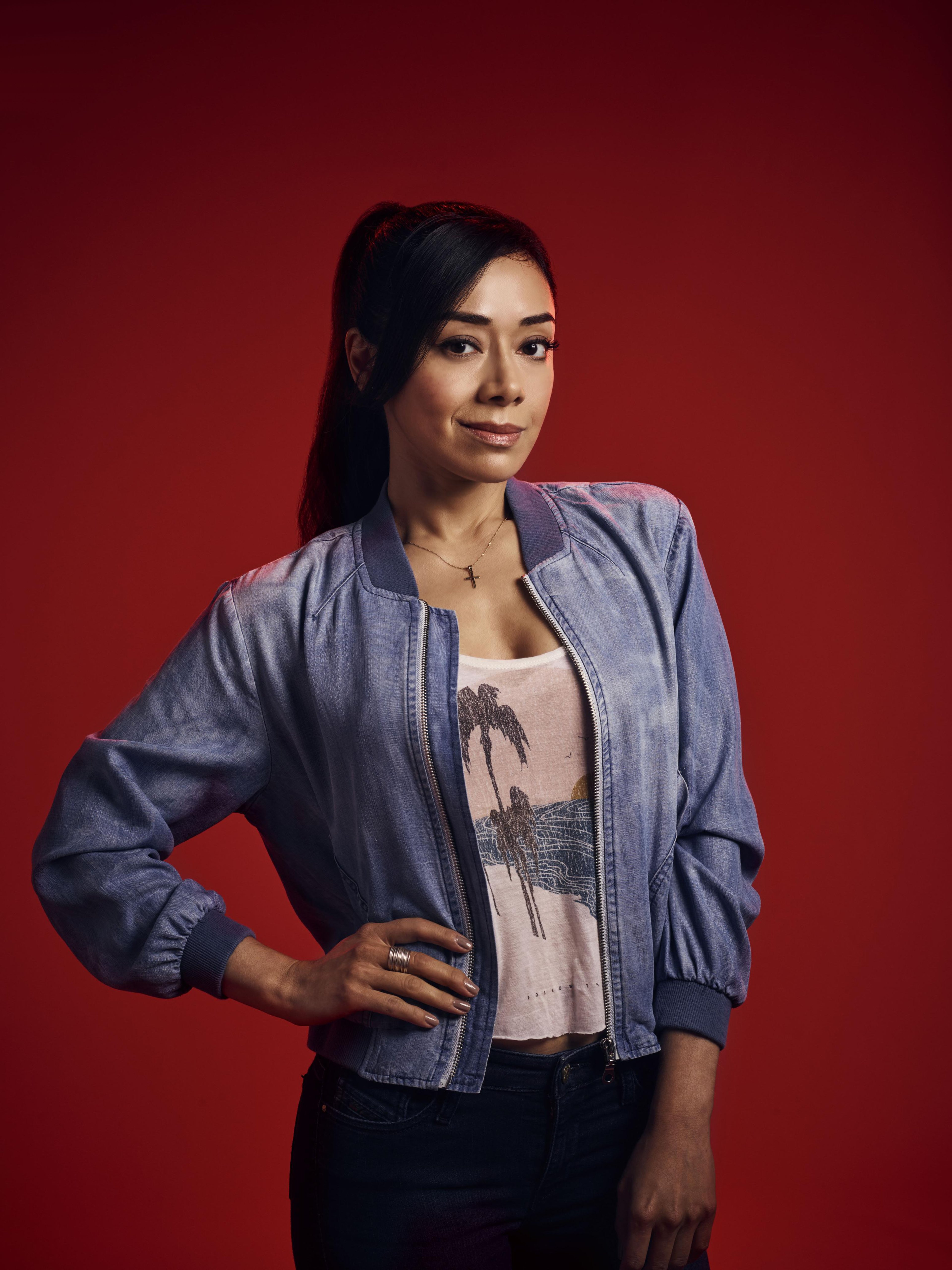 A young woman with dark hair wearing a gray jacket, standing against a red background.