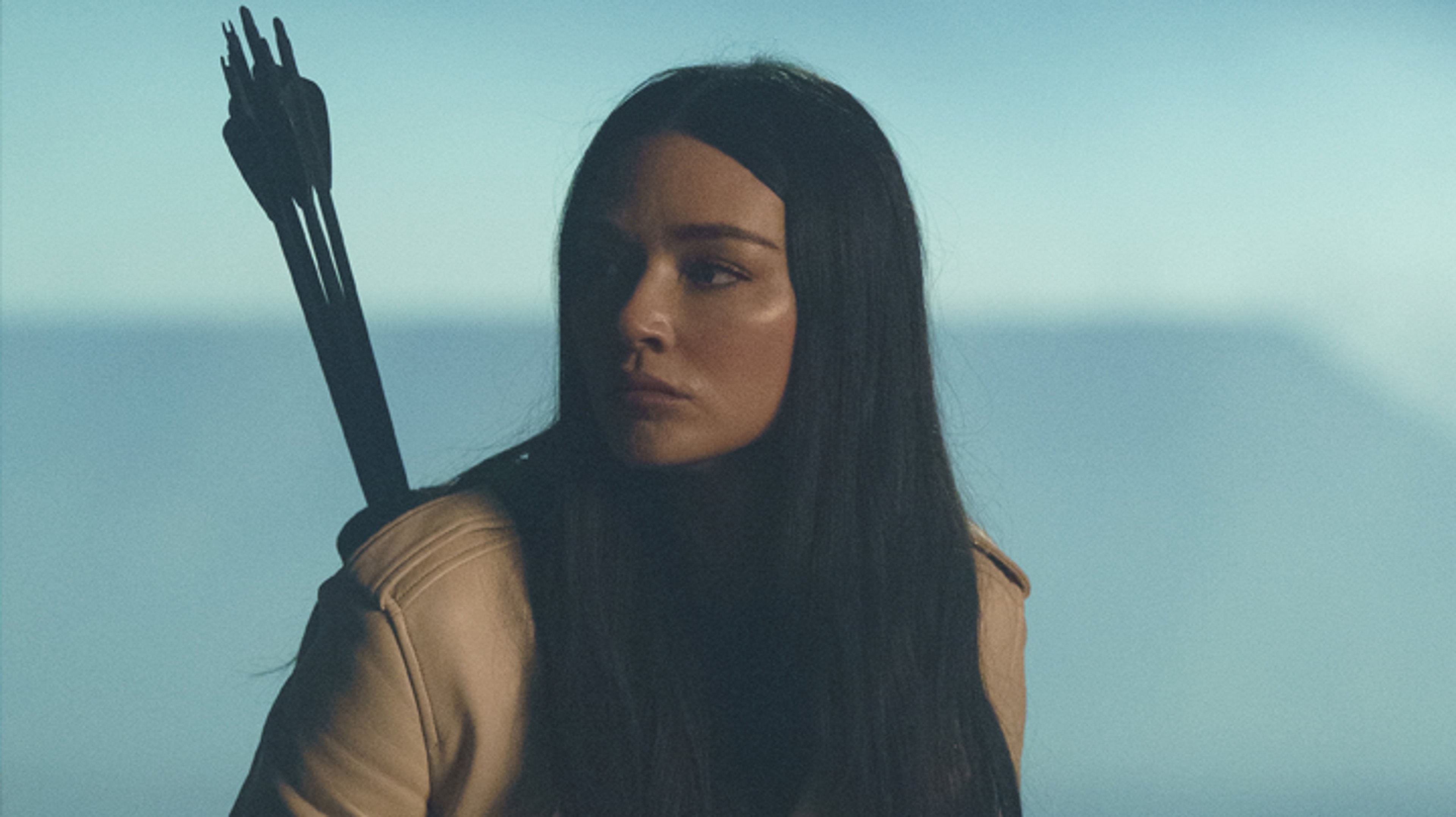 A young woman with long dark hair holding a bow and arrow in an outdoor setting.