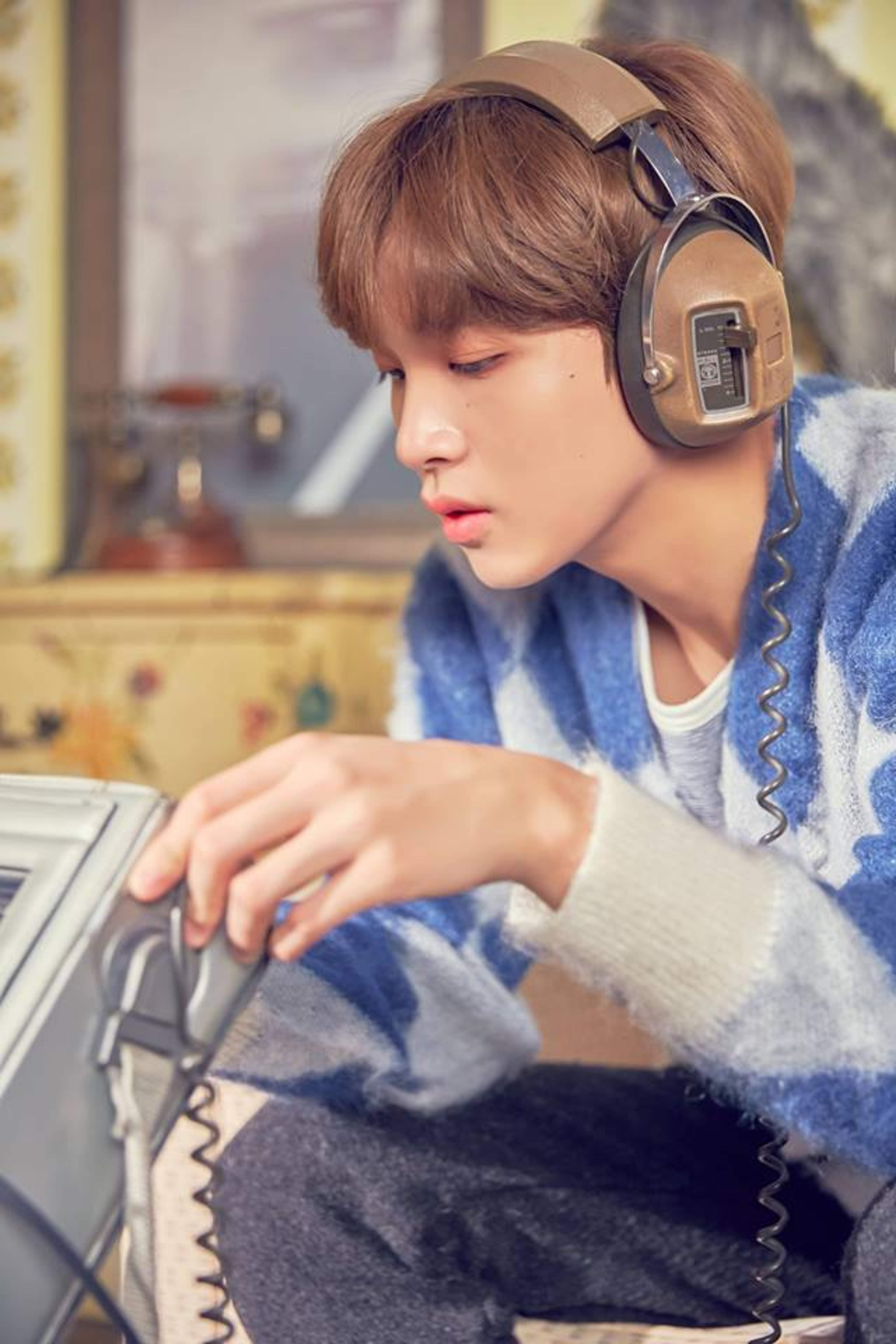 A young man with brown hair wearing headphones and a blue denim jacket, sitting at a desk or table.
