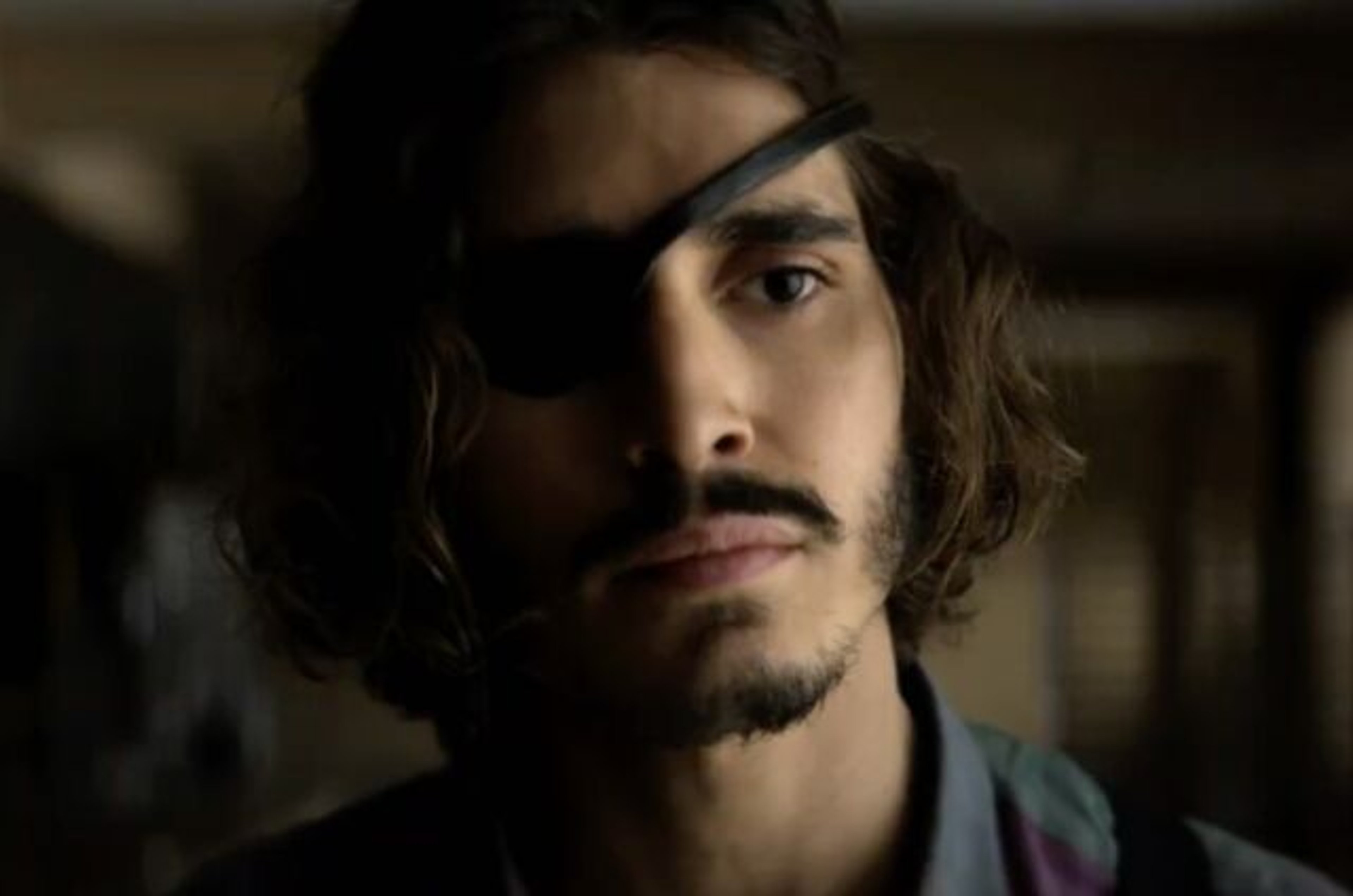 A young man with long dark hair and a beard in a moody, dimly lit setting.