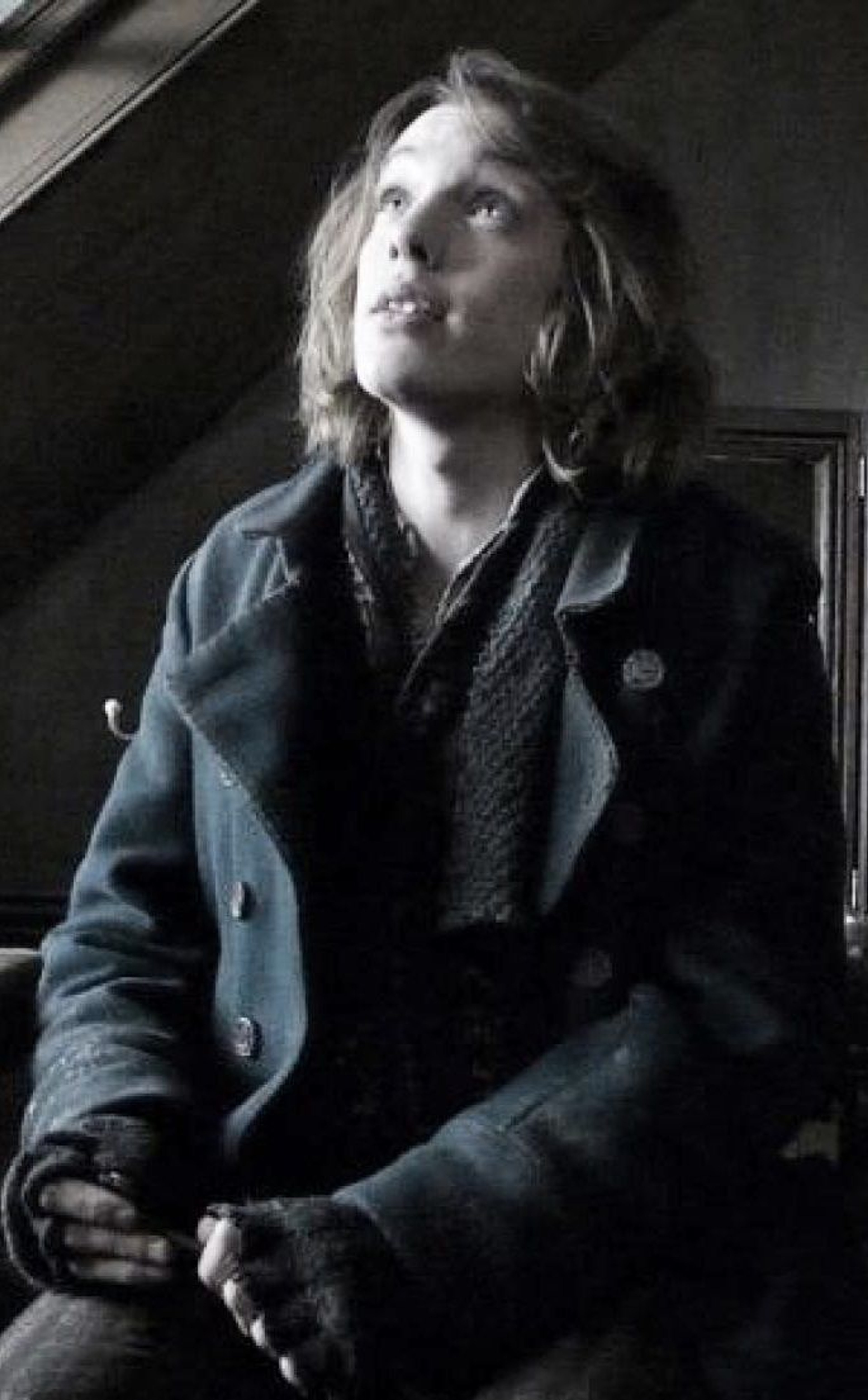 A young man with blonde hair and blue eyes, wearing a dark coat and scarf, gazing off to the side with a thoughtful expression.