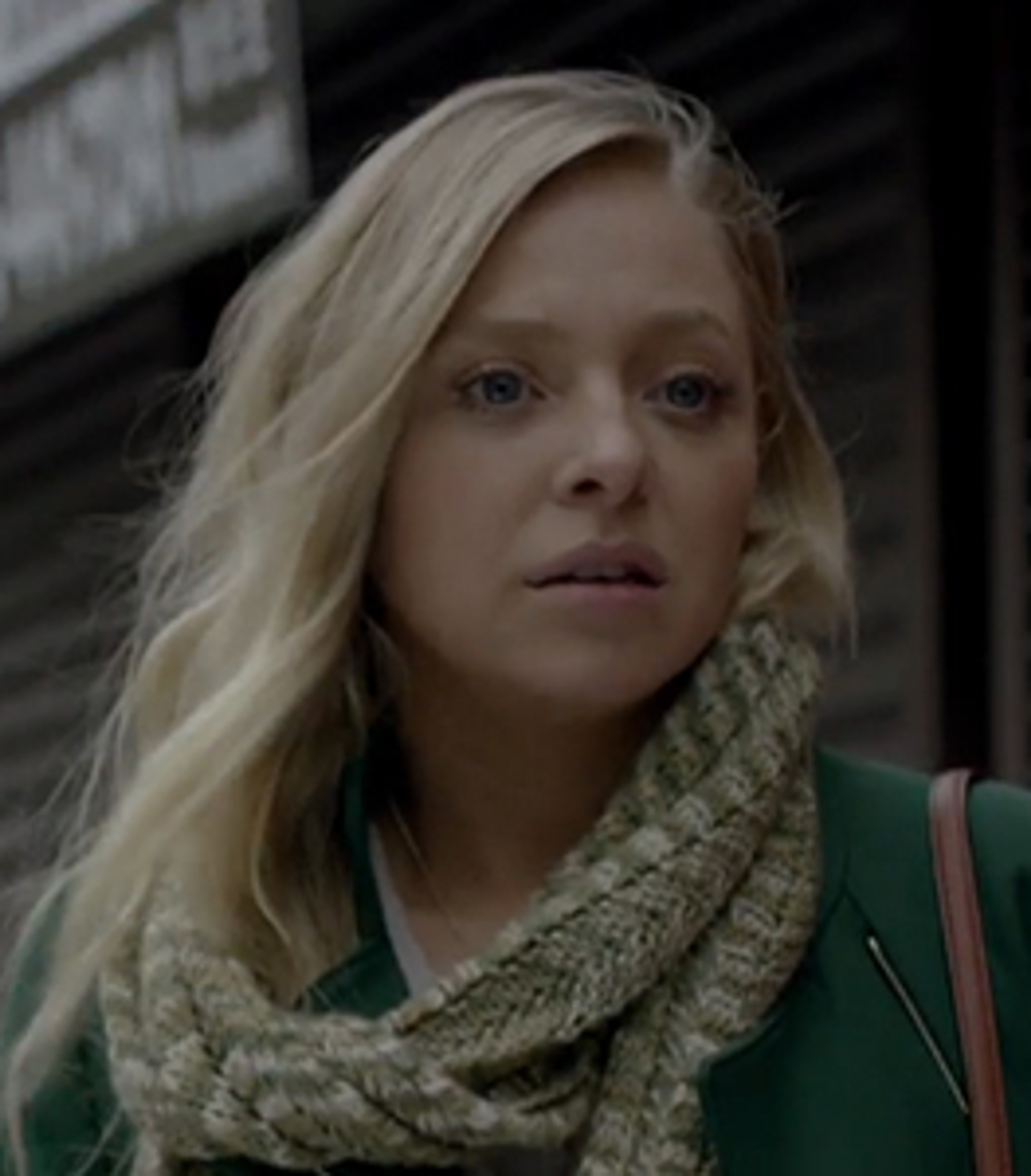 A young woman with blonde hair and blue eyes wearing a green coat and scarf in an urban setting.