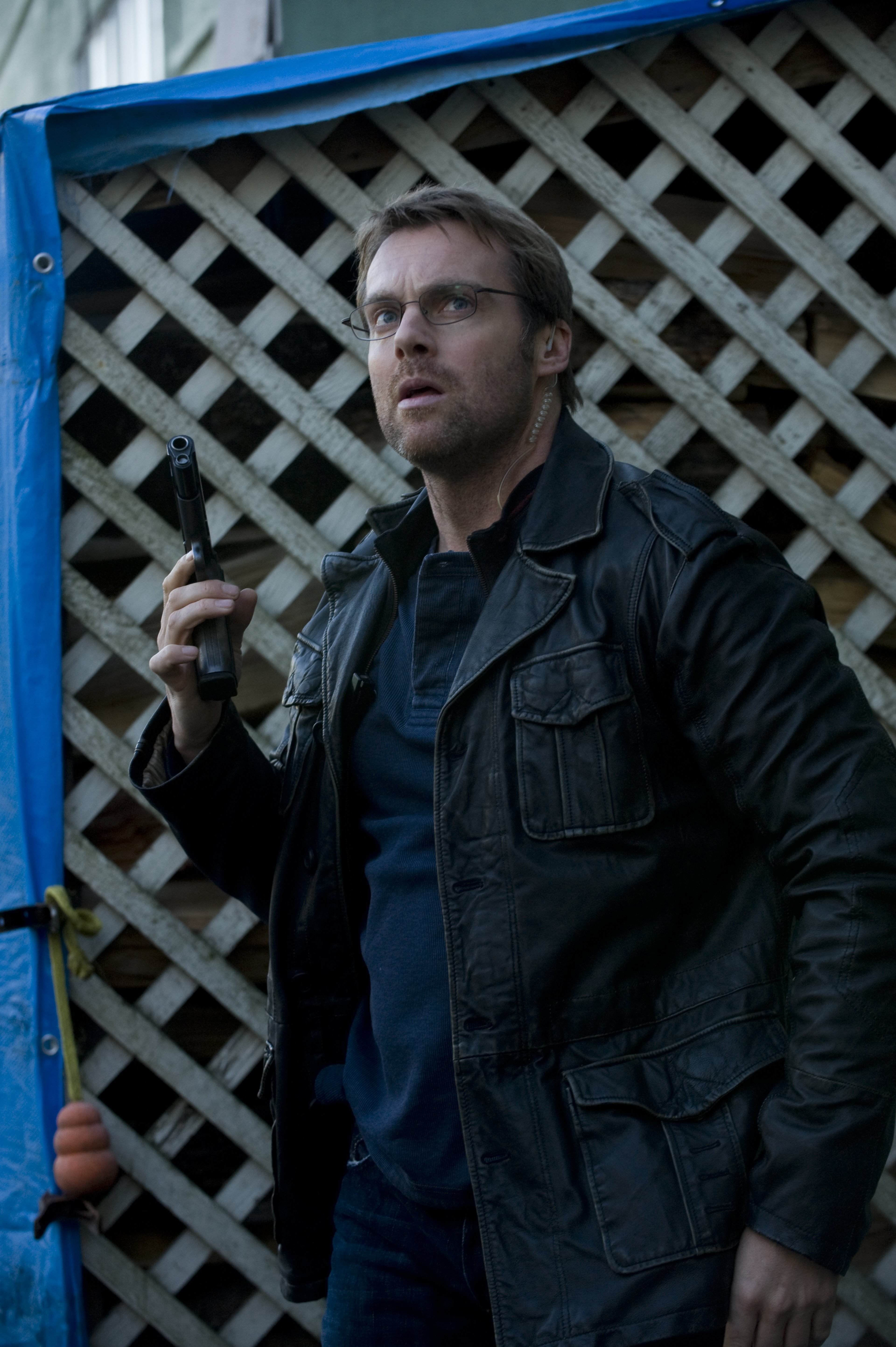 A man with short brown hair and a beard, wearing a dark jacket, standing in front of a lattice fence or gate.