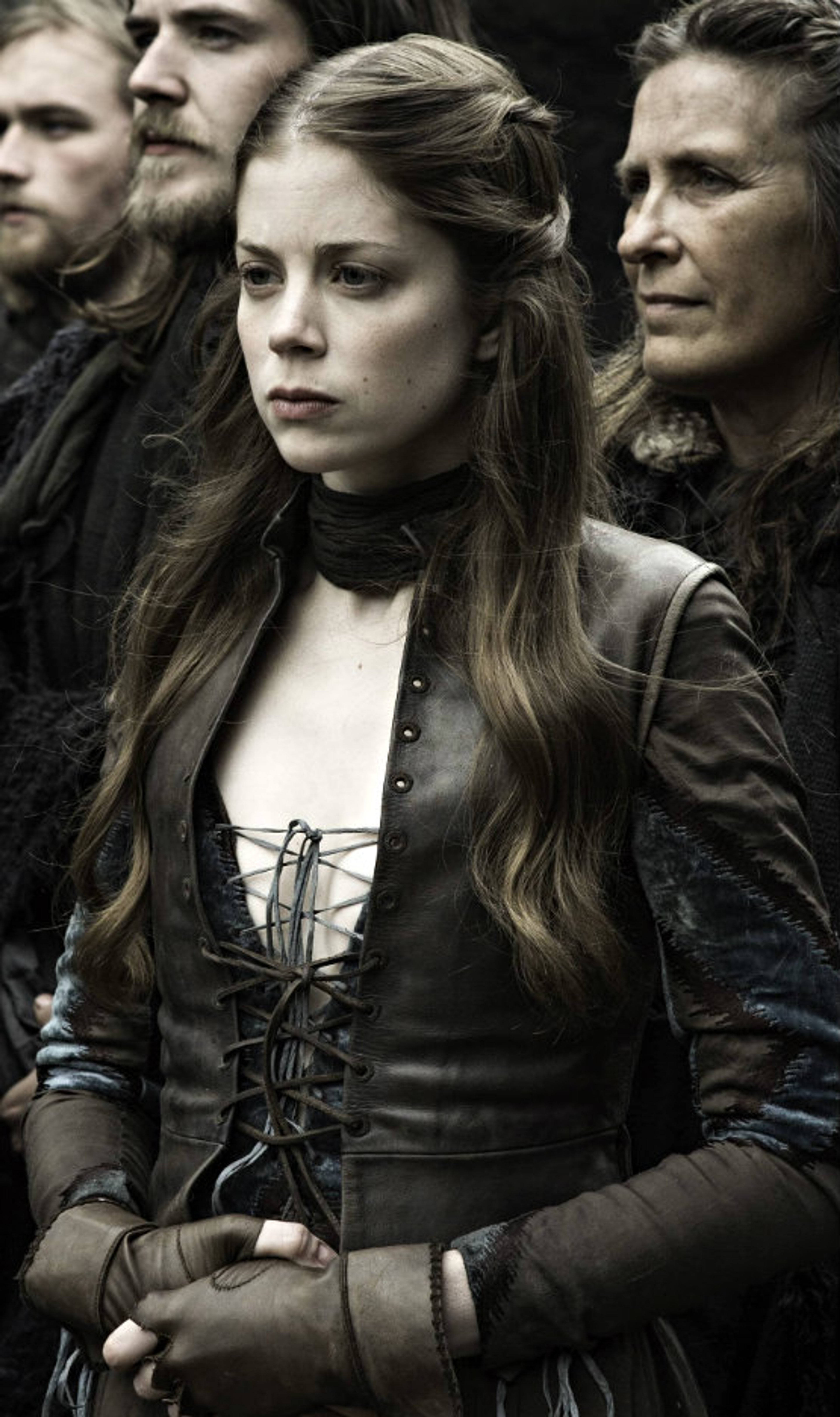 A young woman in a dark leather outfit with a serious expression, surrounded by other people in a medieval or fantasy setting.