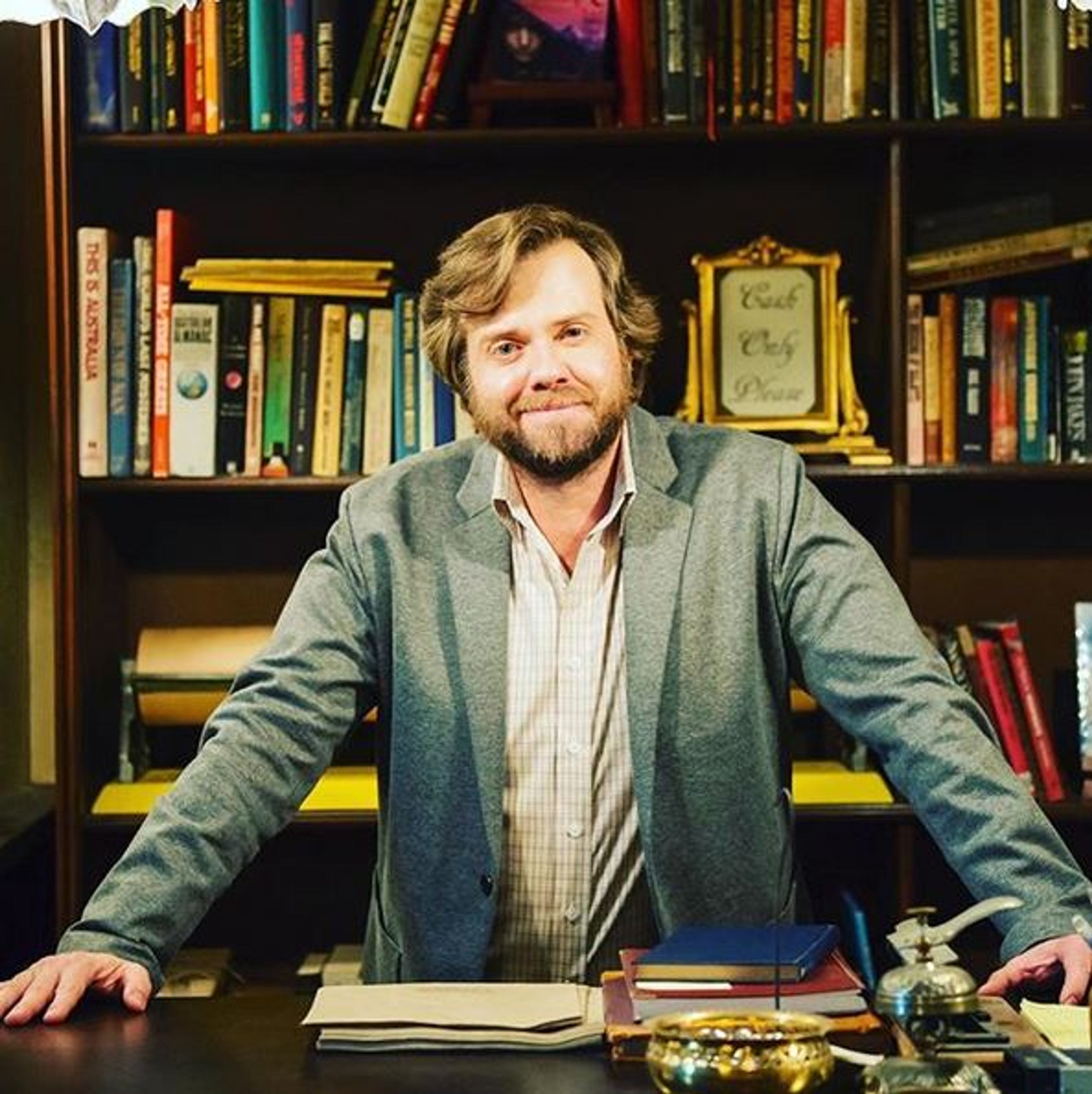 A middle-aged man with a beard and glasses, sitting at a desk in a library or study.
