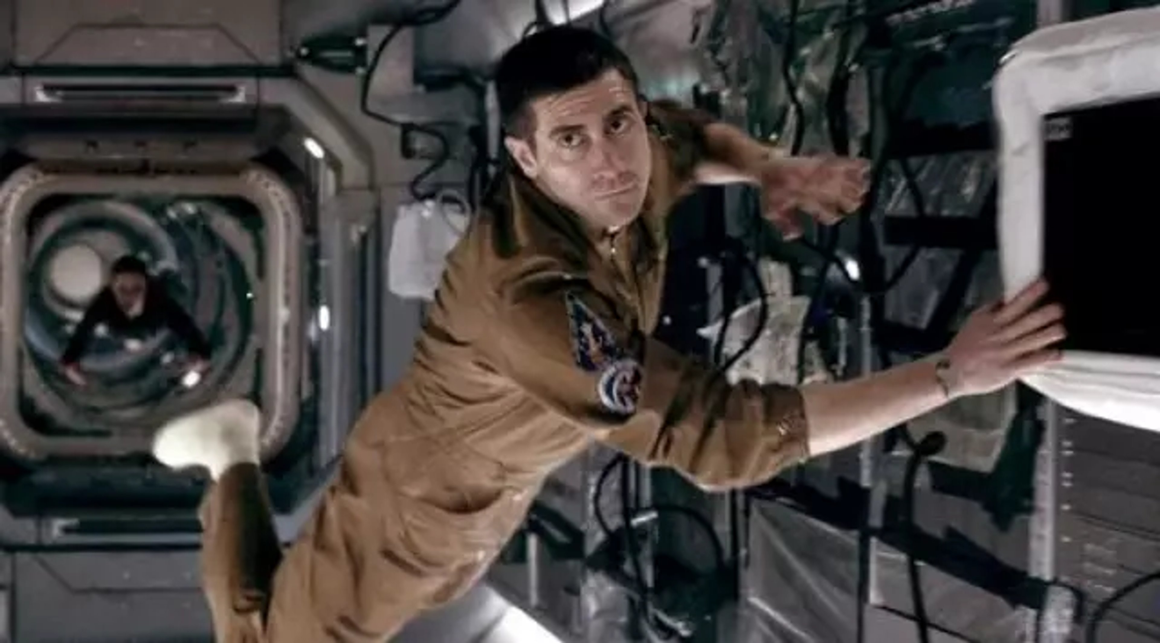 A middle-aged man in a tan jacket sitting at a control panel in a spacecraft or space station