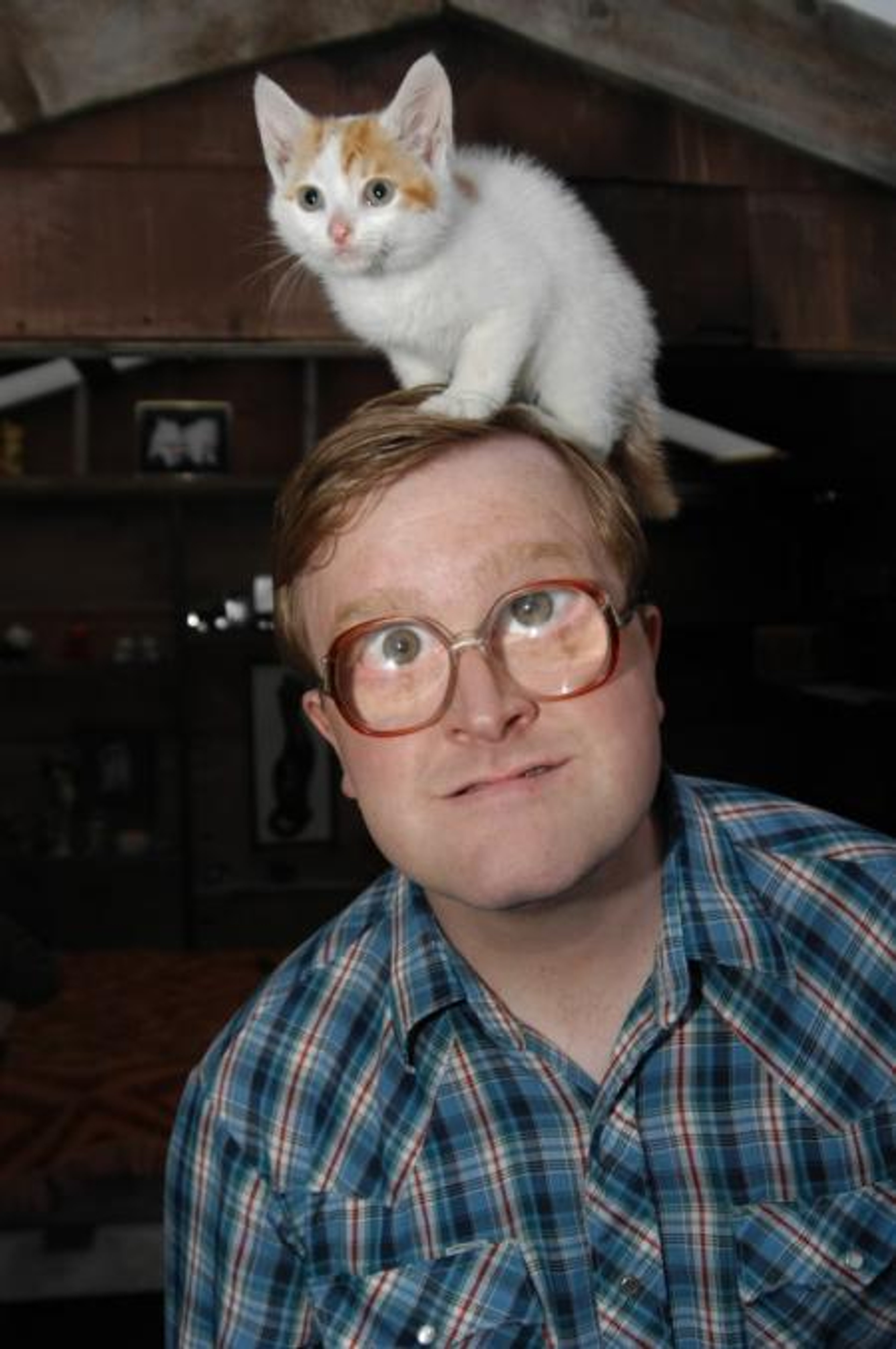 A man with glasses and a plaid shirt, with a small white cat perched on his head.