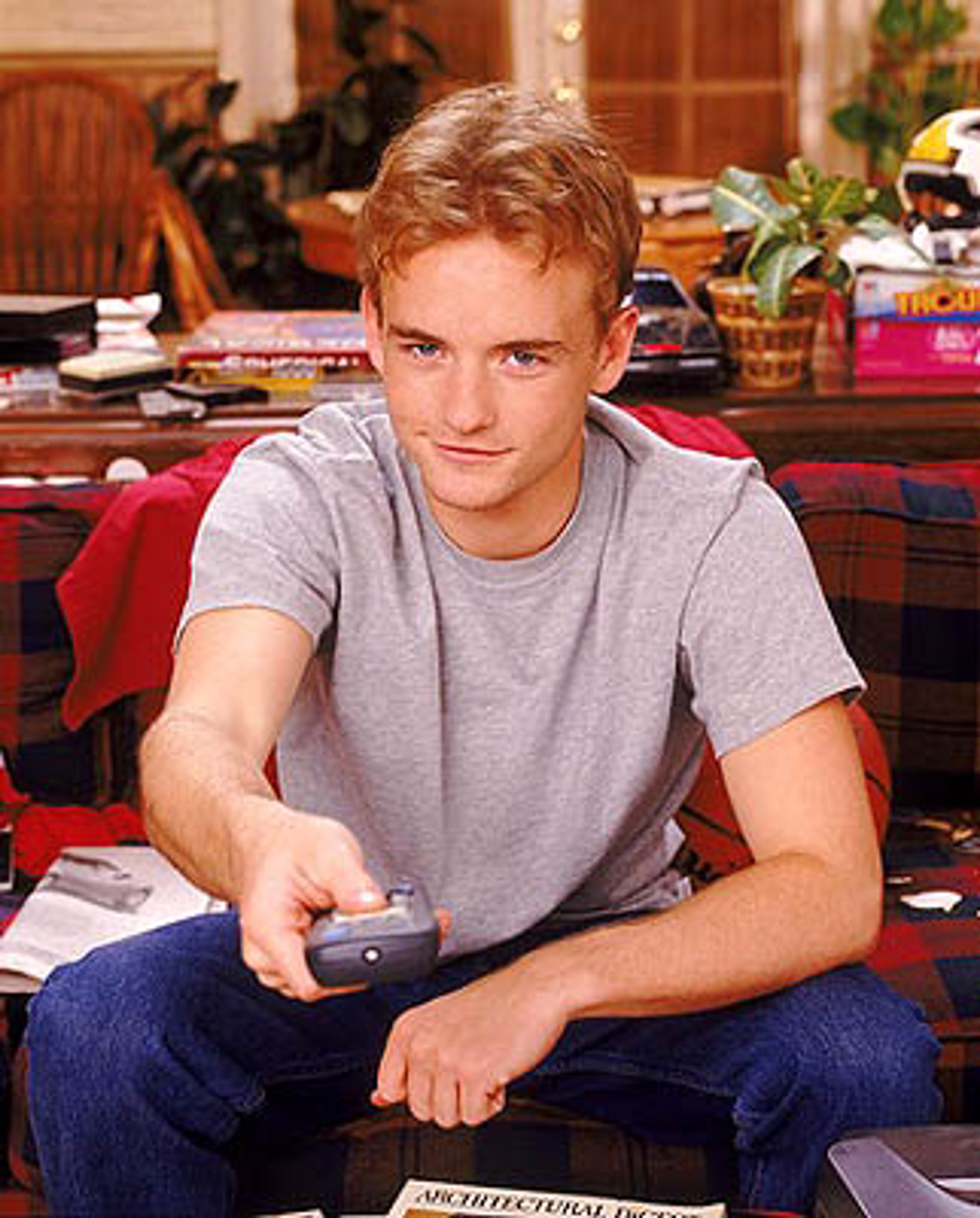 A young man with blond hair sitting in a messy room, holding a game controller or remote.