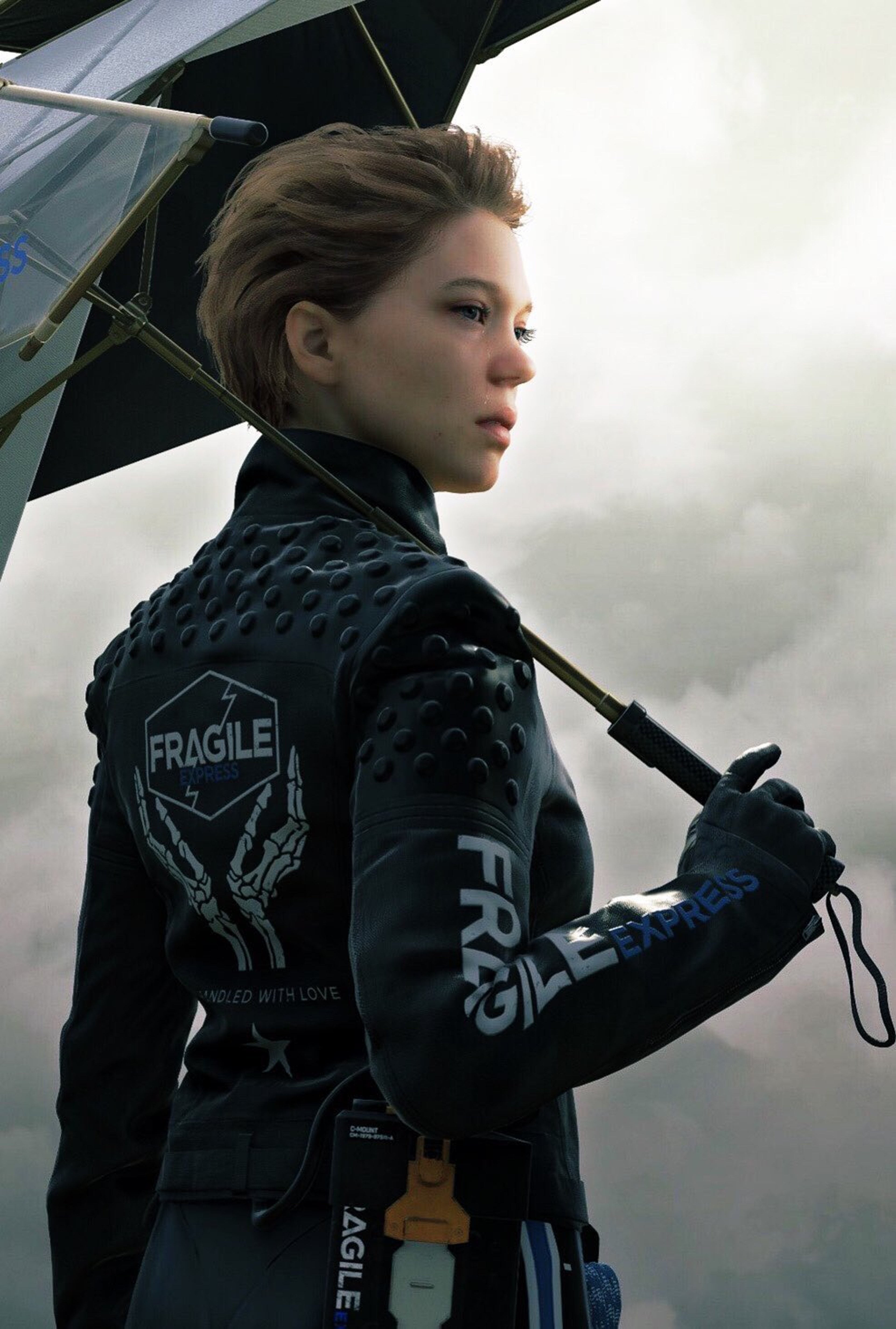 A woman with short blonde hair wearing a black jacket with the word 'FRAGILE' printed on it, holding an umbrella and looking off to the side.
