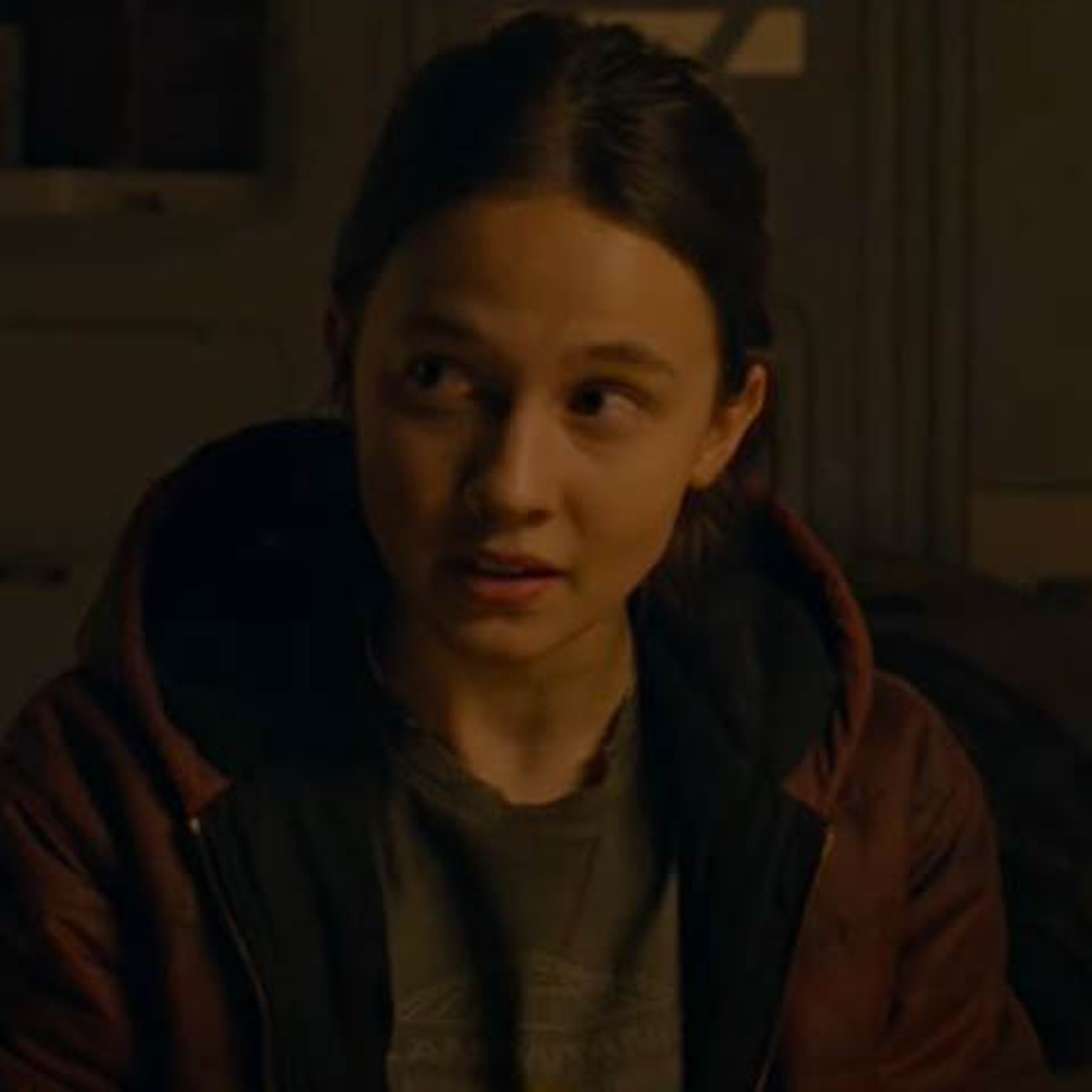 A young woman with dark hair wearing a red jacket in a dimly lit indoor setting.