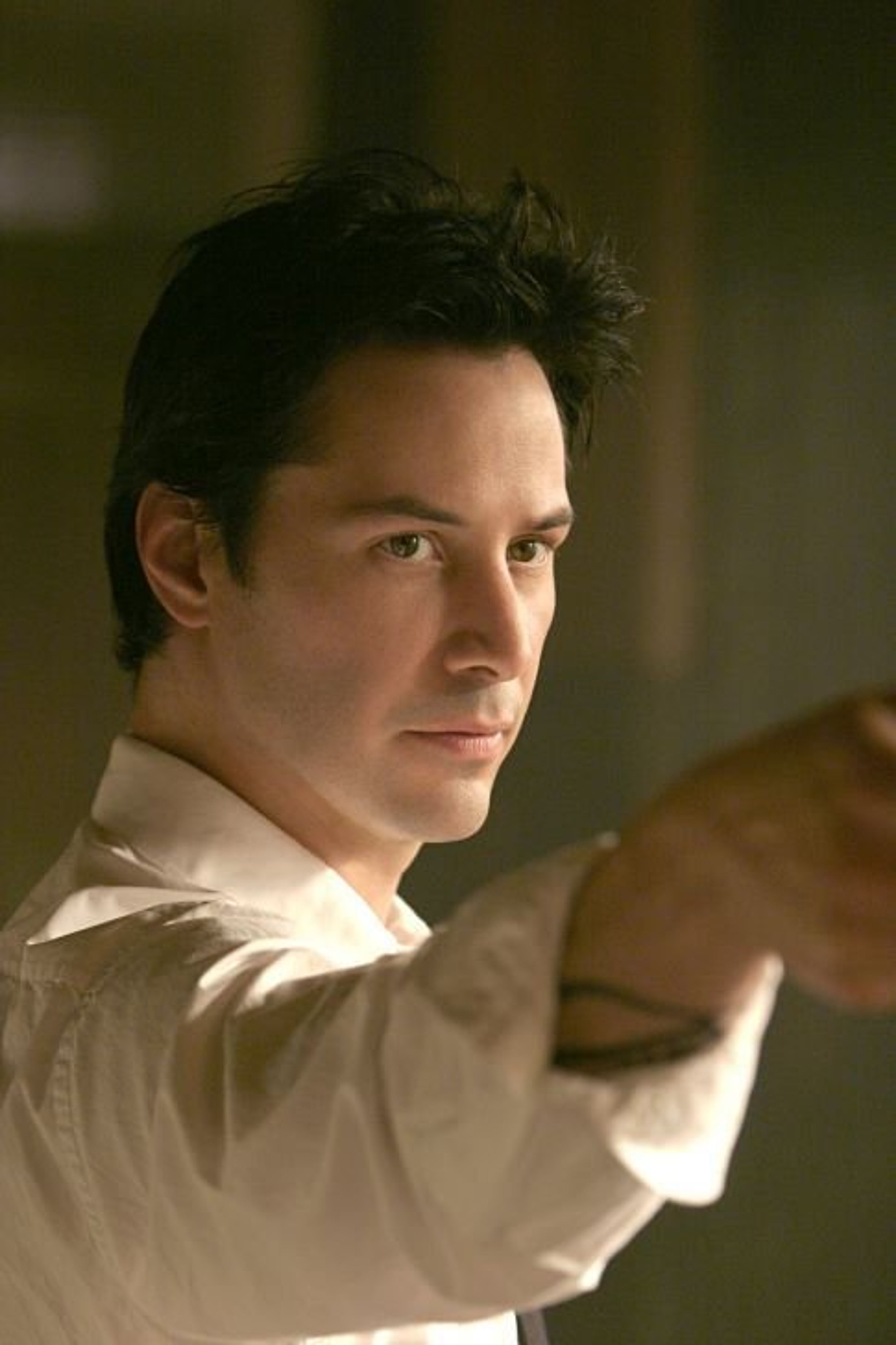 A man with dark hair and a serious expression, wearing a white shirt in a dimly lit room.