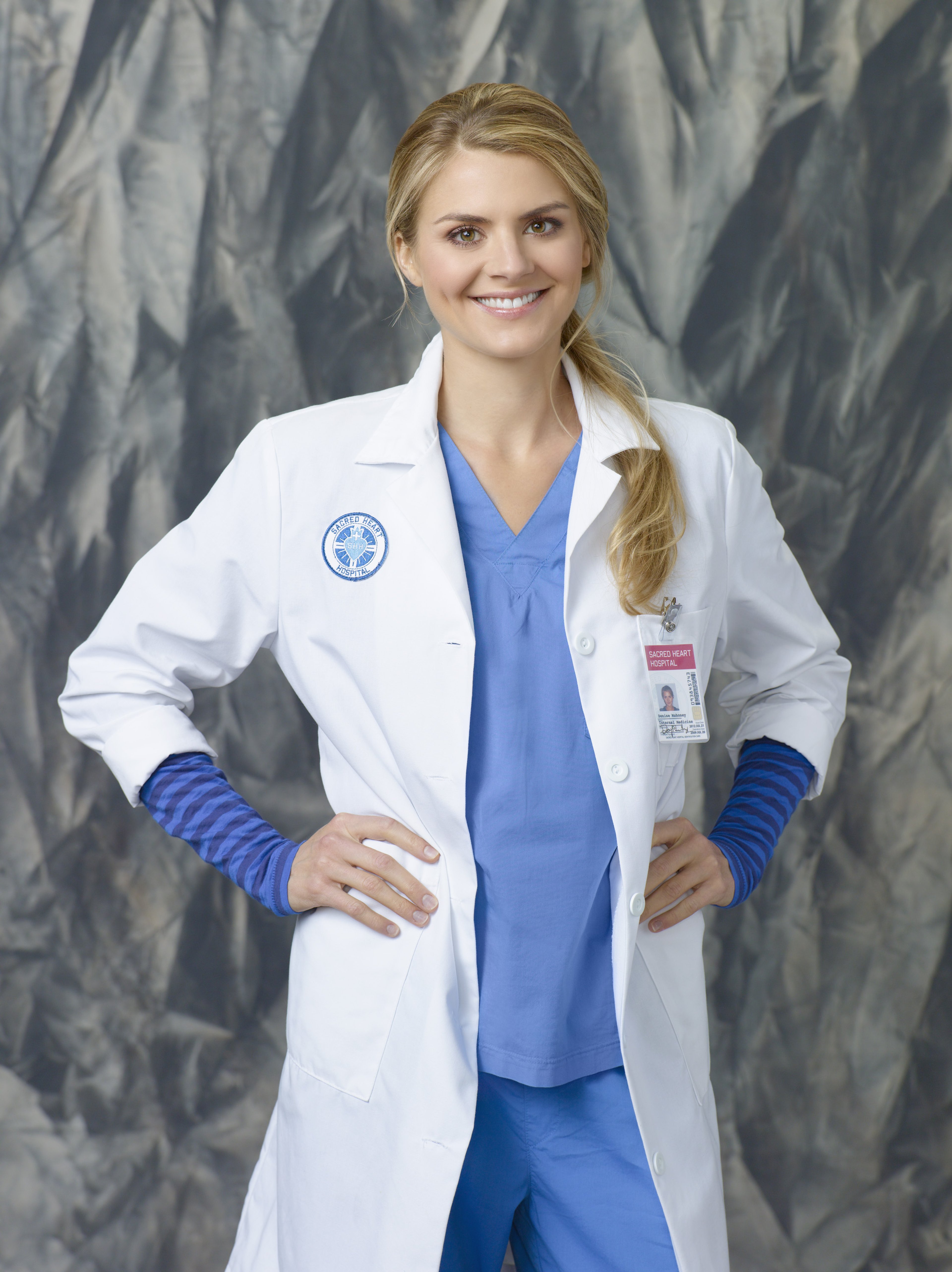 A smiling blonde woman wearing a white doctor's coat and blue scrubs