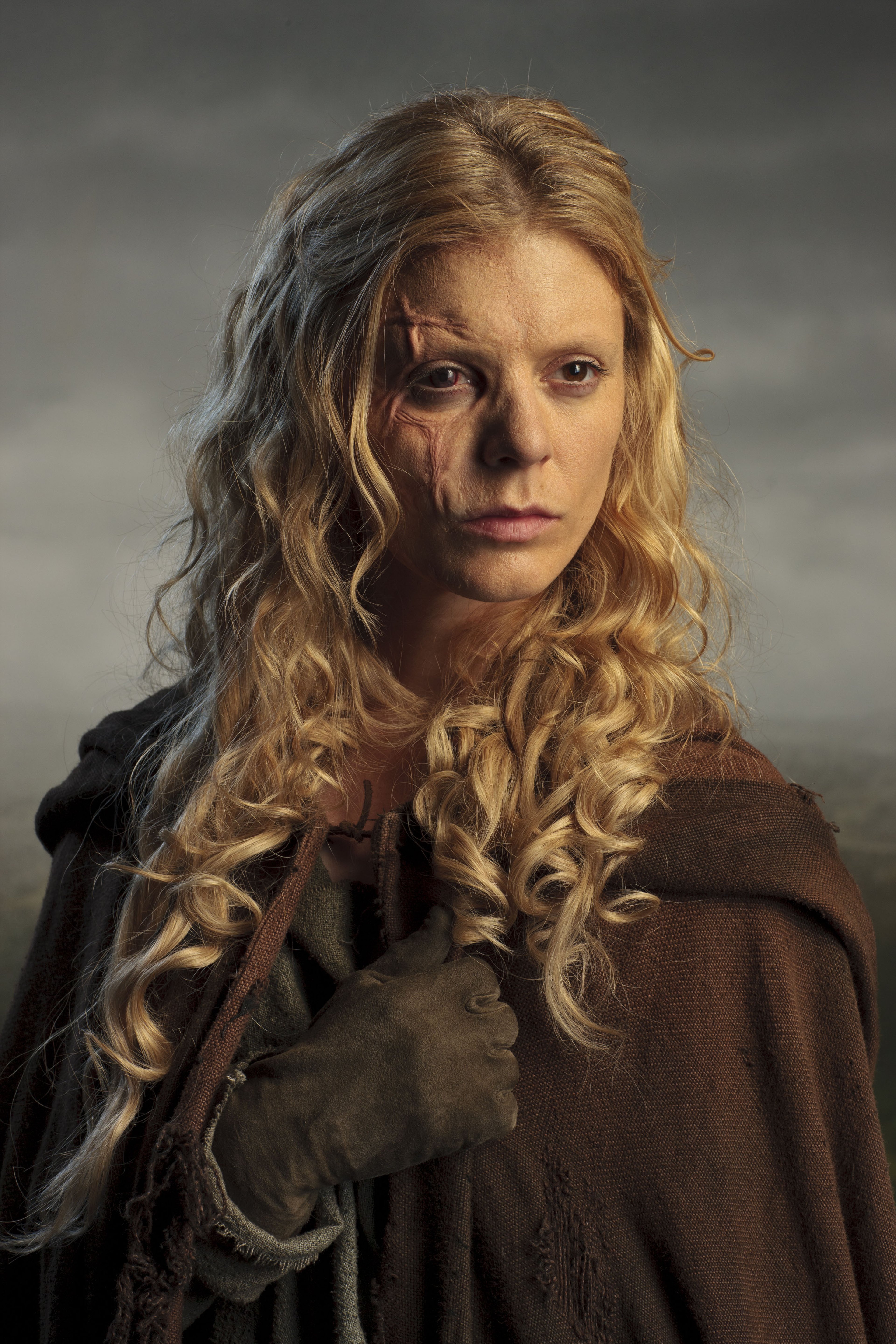 A woman with long, curly blonde hair wearing a dark brown cloak, with a serious expression on her face.