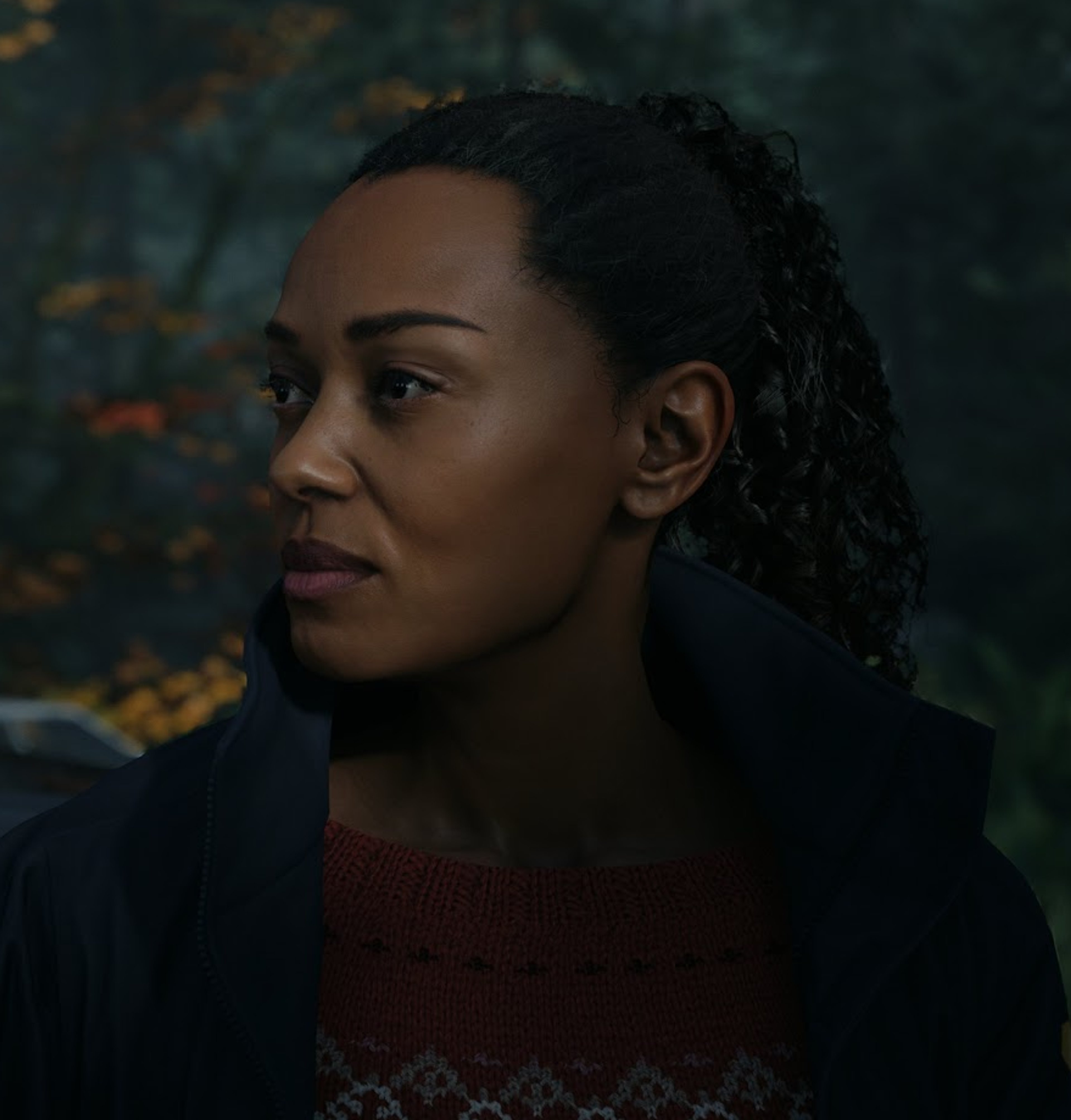 A young woman with dark skin and curly hair in a serious expression, wearing a red shirt and dark jacket in a natural outdoor setting.