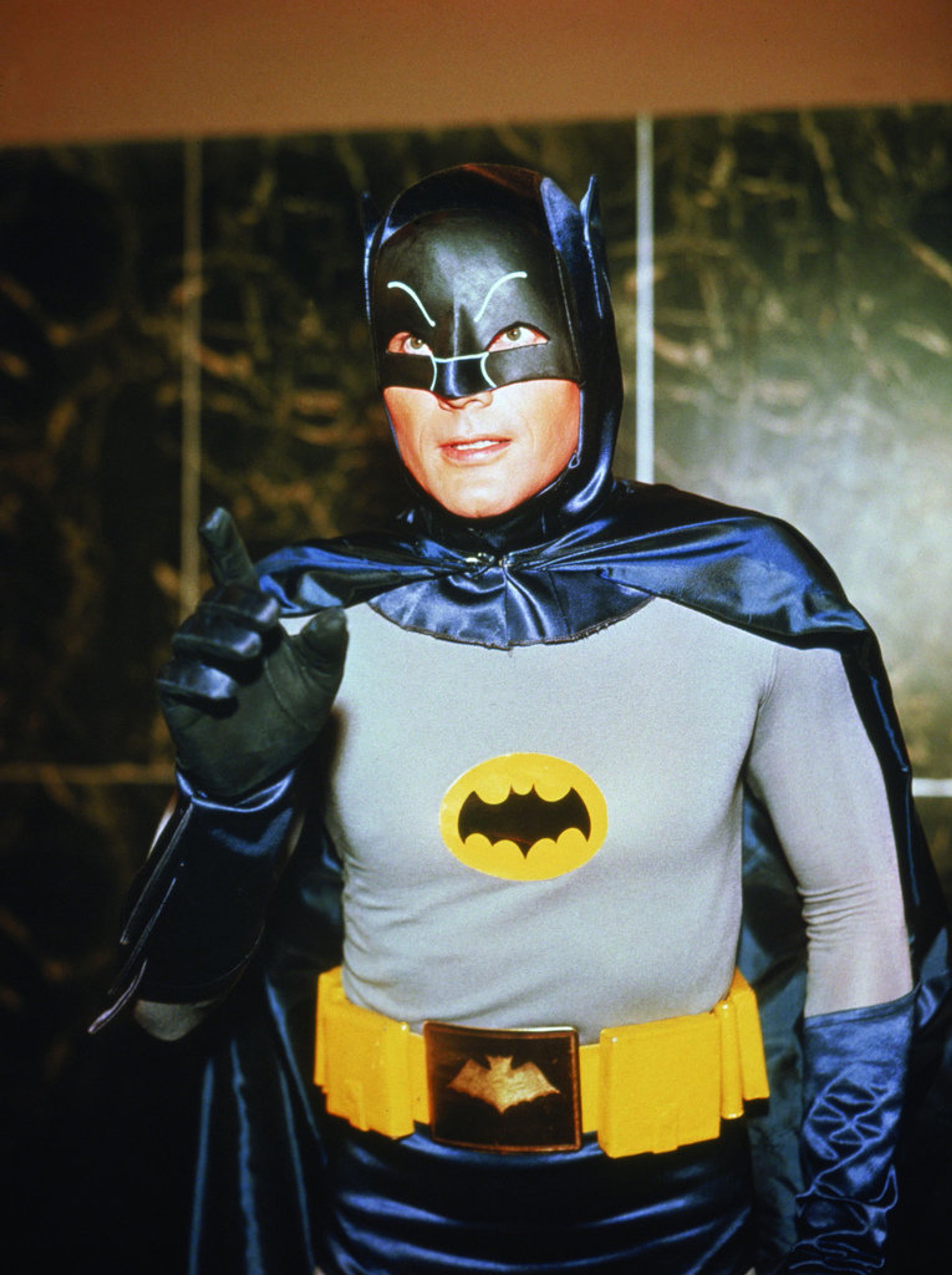 A man dressed in a classic 1960s Batman costume, standing in a dark, shadowy environment.