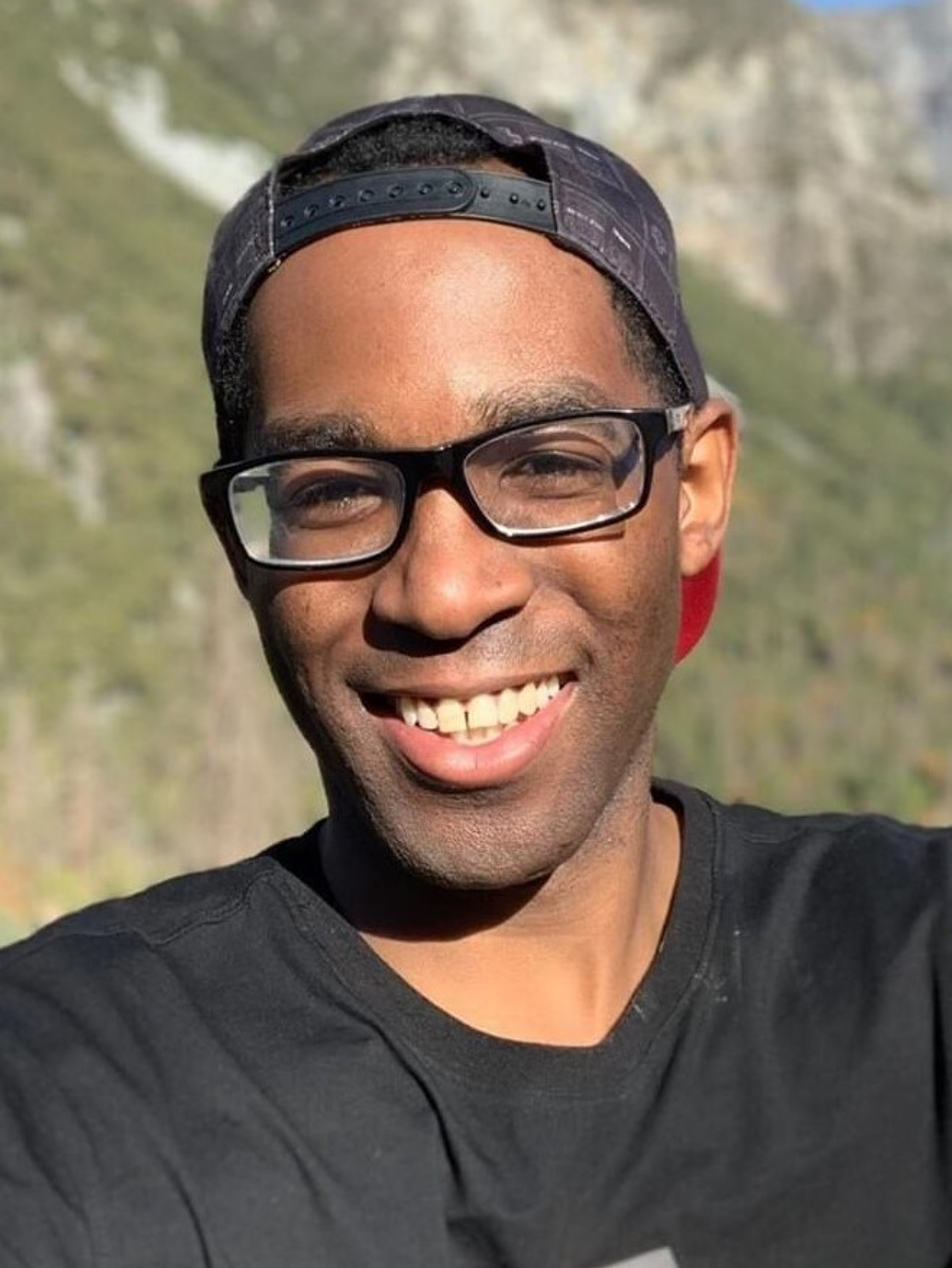 A smiling Black man wearing glasses and a black hat