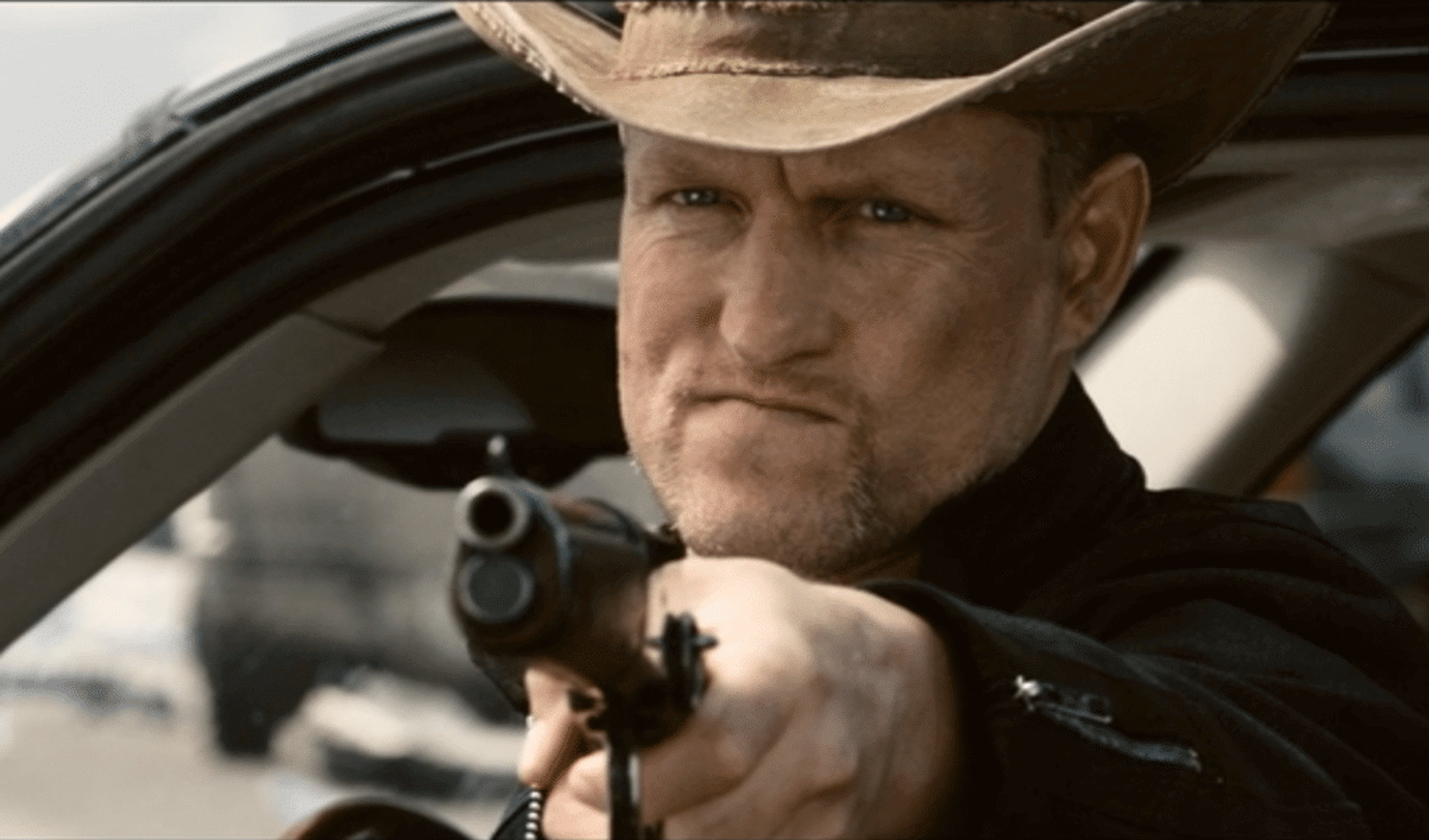 A middle-aged man with a bald head wearing a cowboy hat, jeans, and a leather jacket, holding a gun with a serious expression.