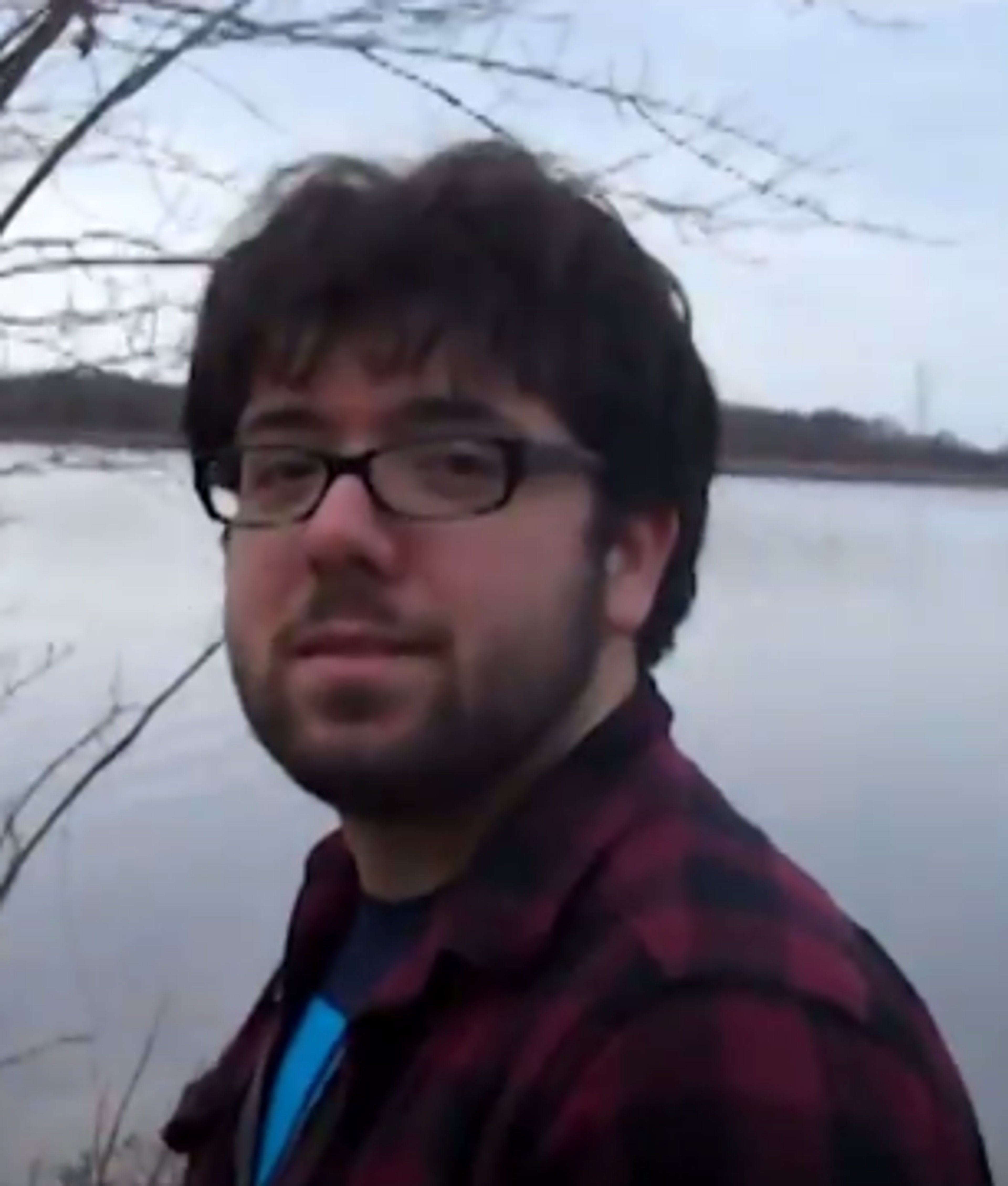 A man with dark hair and glasses wearing a plaid shirt, standing outdoors near a body of water.