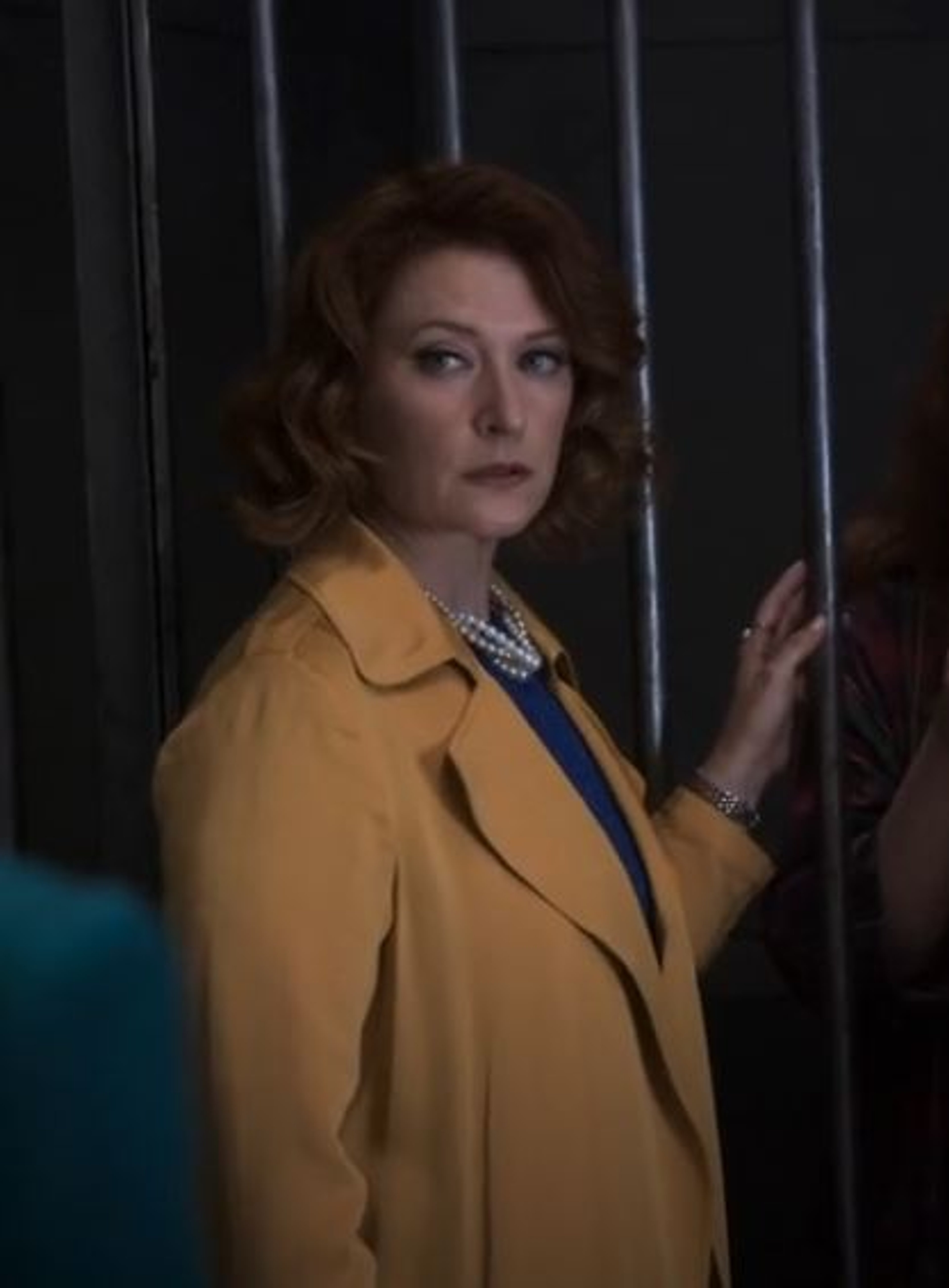 A woman with curly red hair wearing a yellow coat standing in front of a dark background with vertical bars