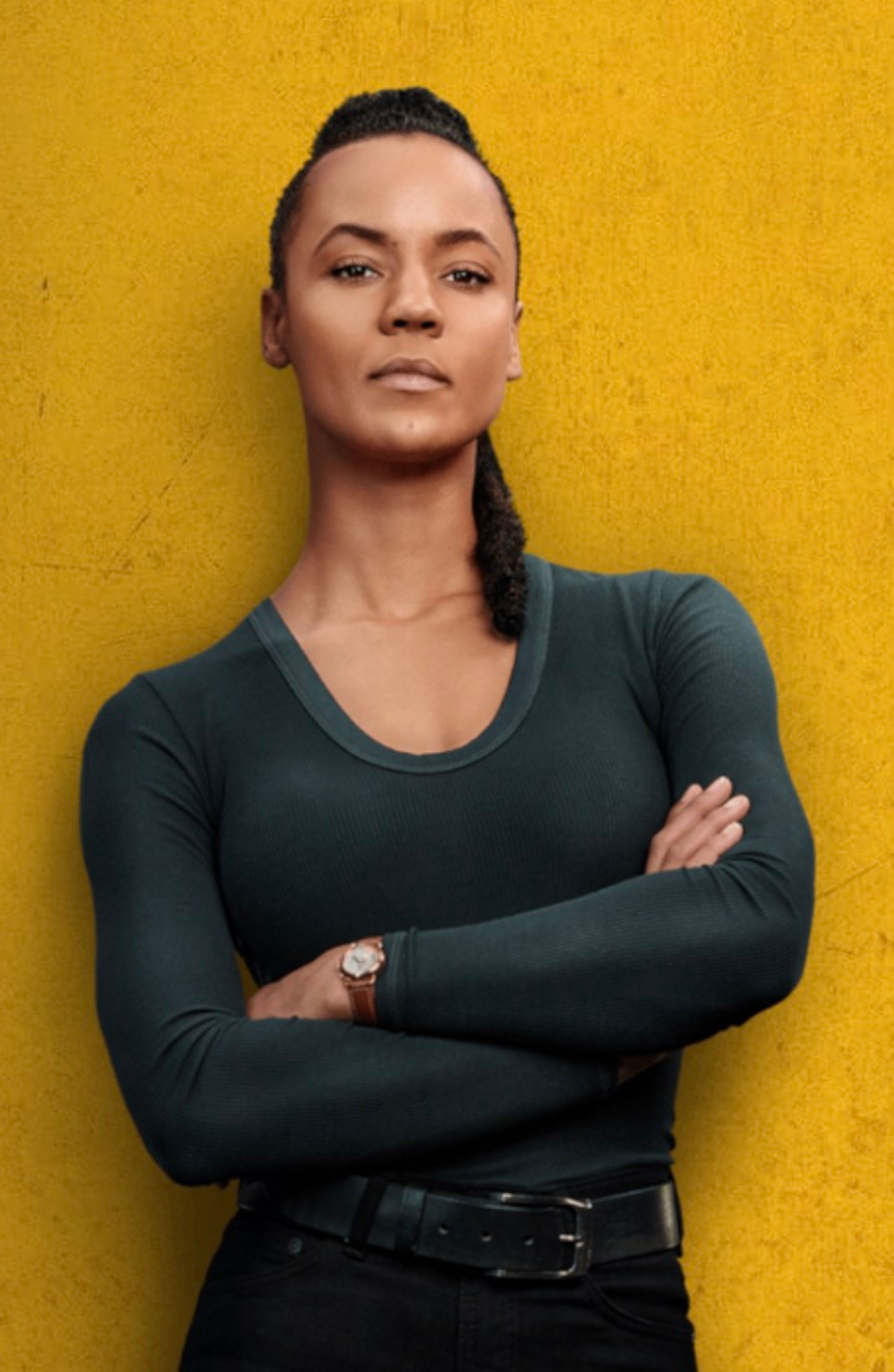 A woman with braided hair standing against a yellow wall, with a serious expression on her face.