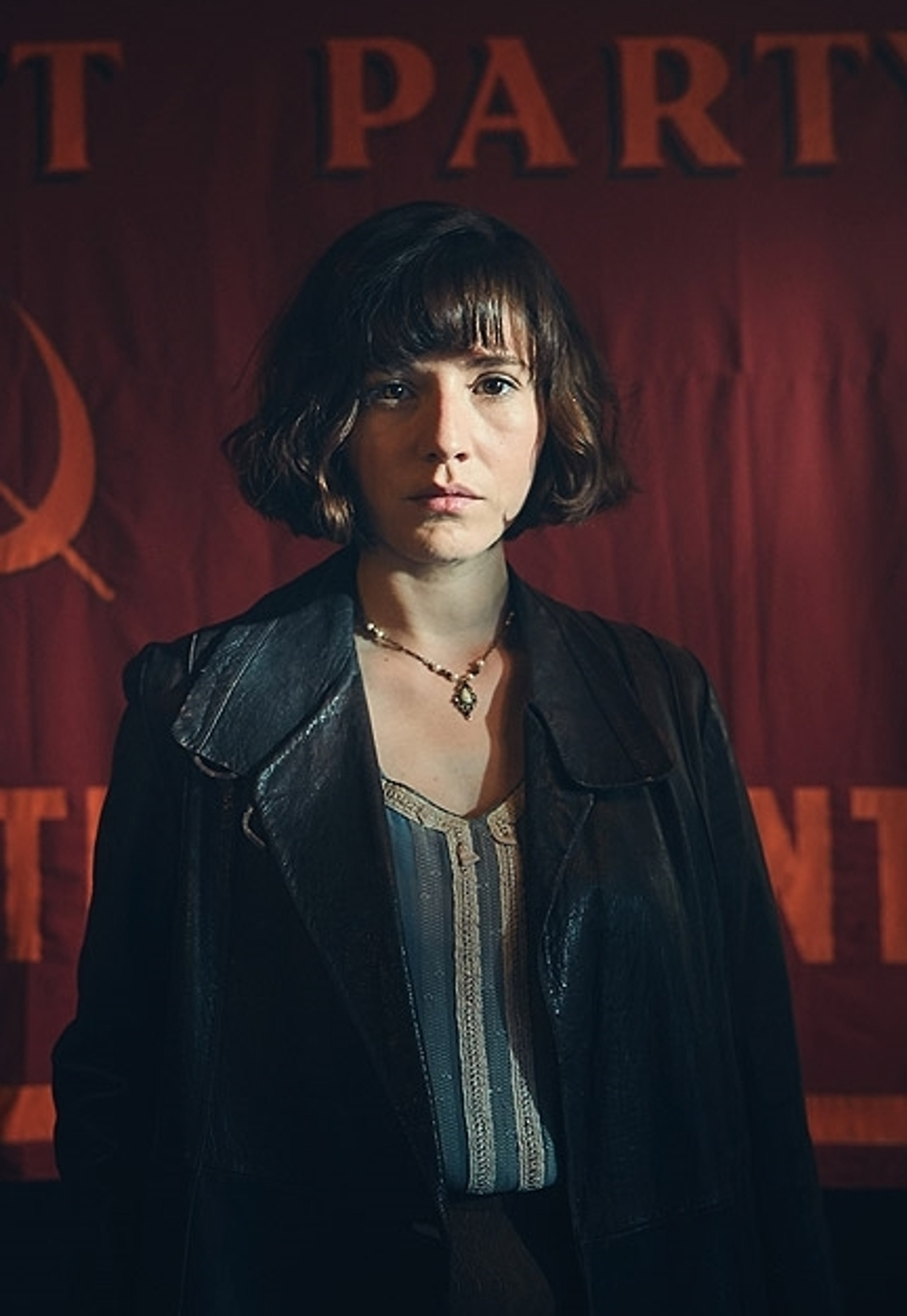 A young woman with short dark hair wearing a black jacket, standing in front of a red background.
