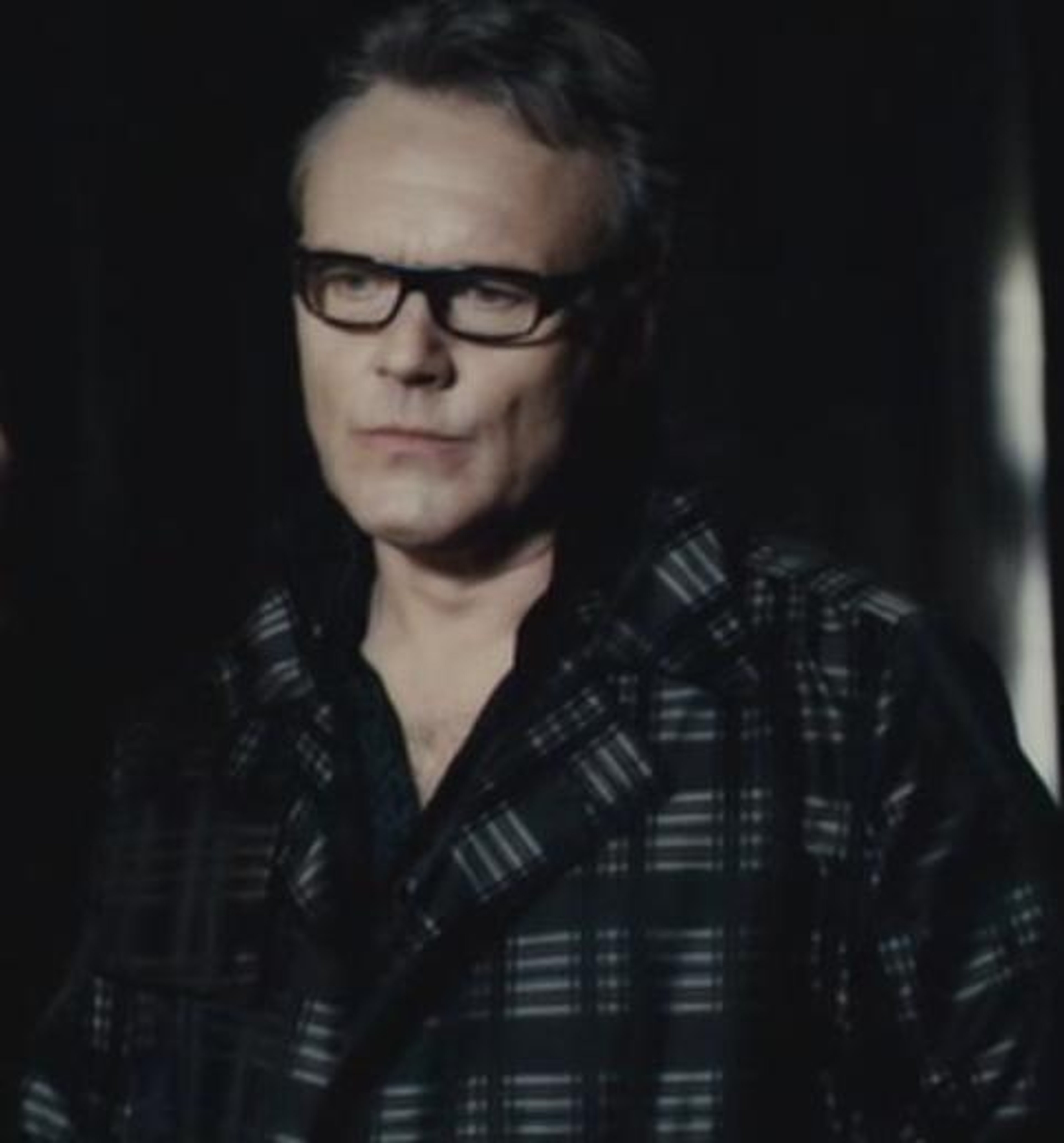 A middle-aged man with greying hair and glasses, wearing a plaid shirt and having a serious expression.