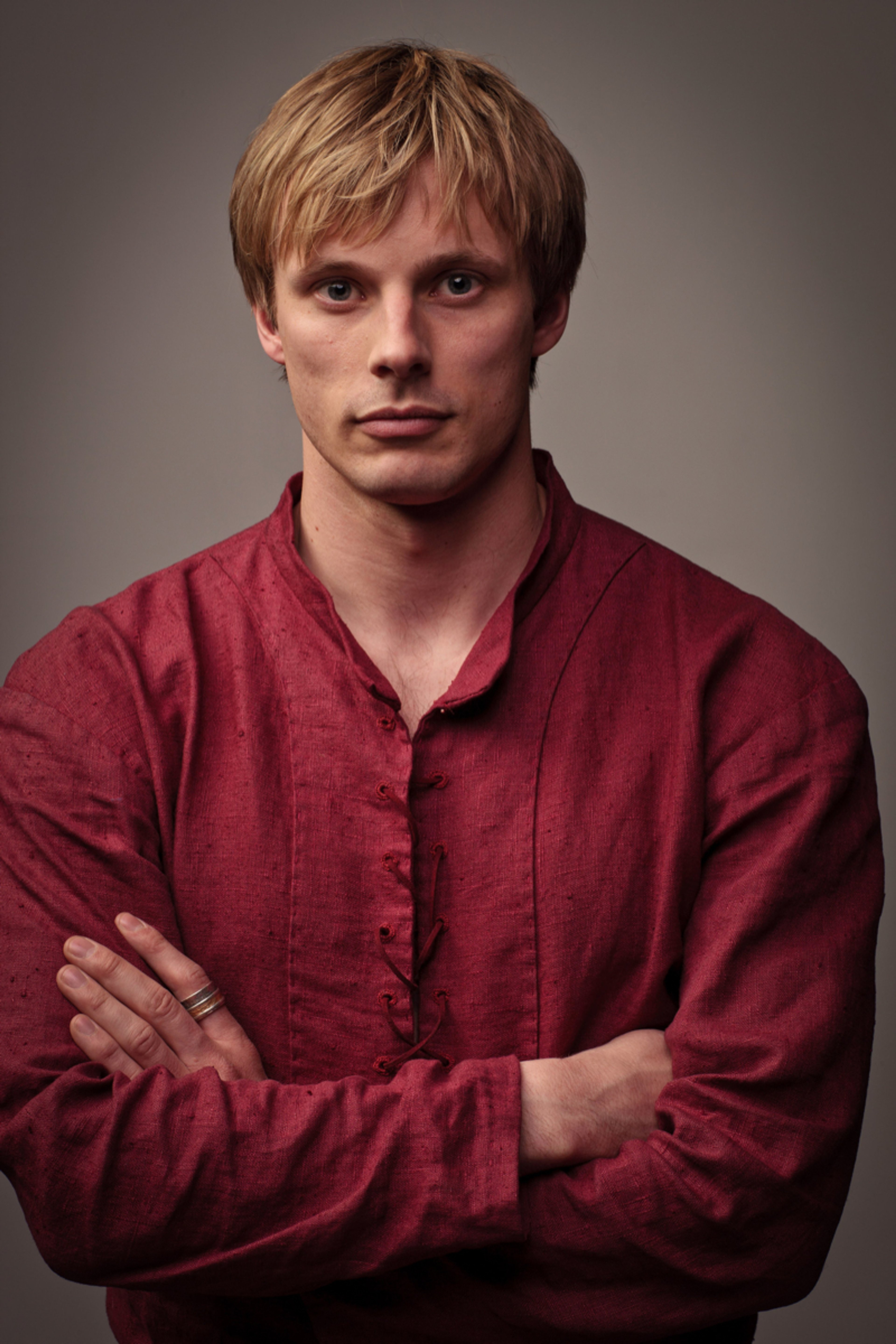 A young man with short blonde hair wearing a red shirt, with a serious expression on his face and his arms crossed.