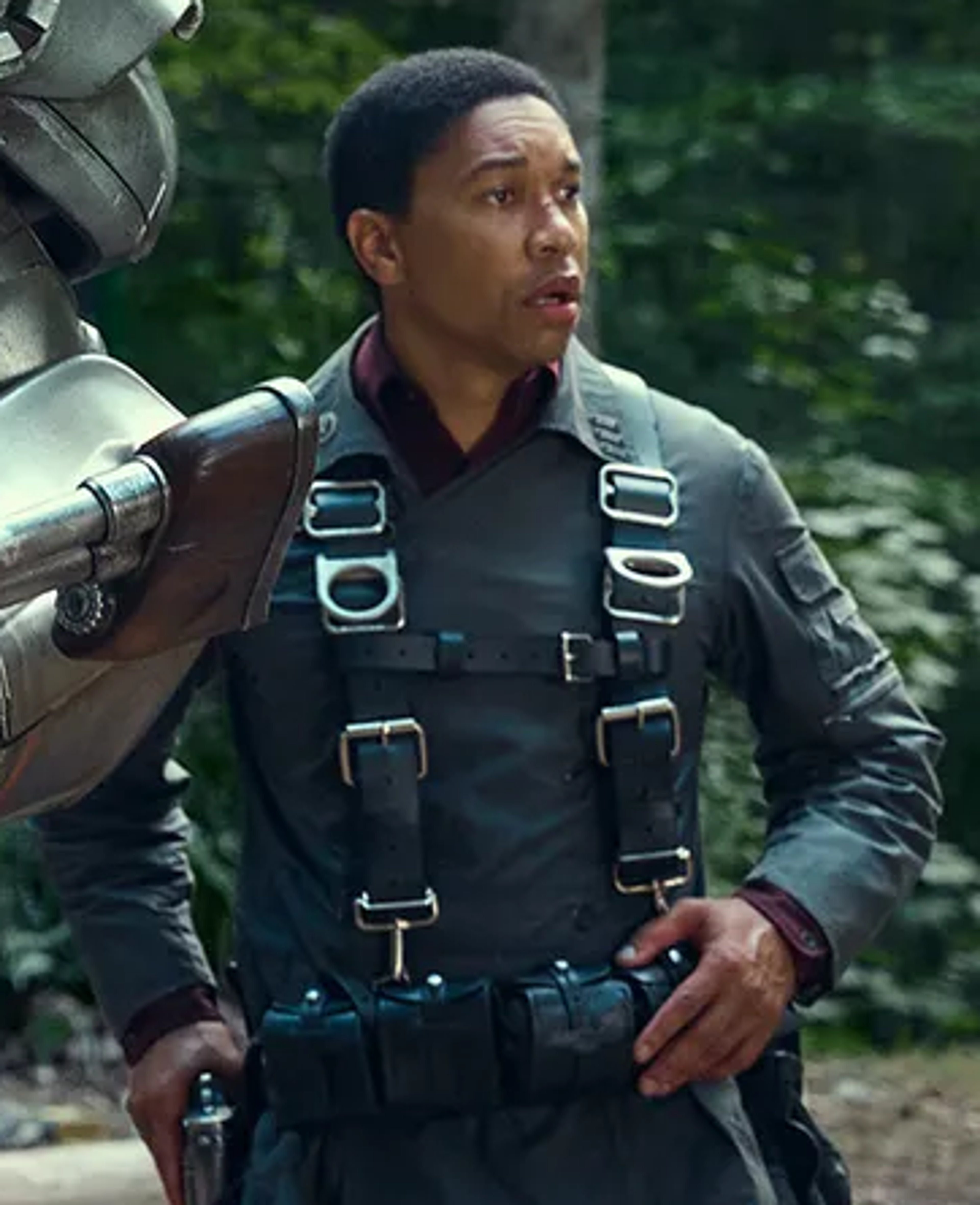 A young Black man in a military-style uniform standing in a forested outdoor setting.