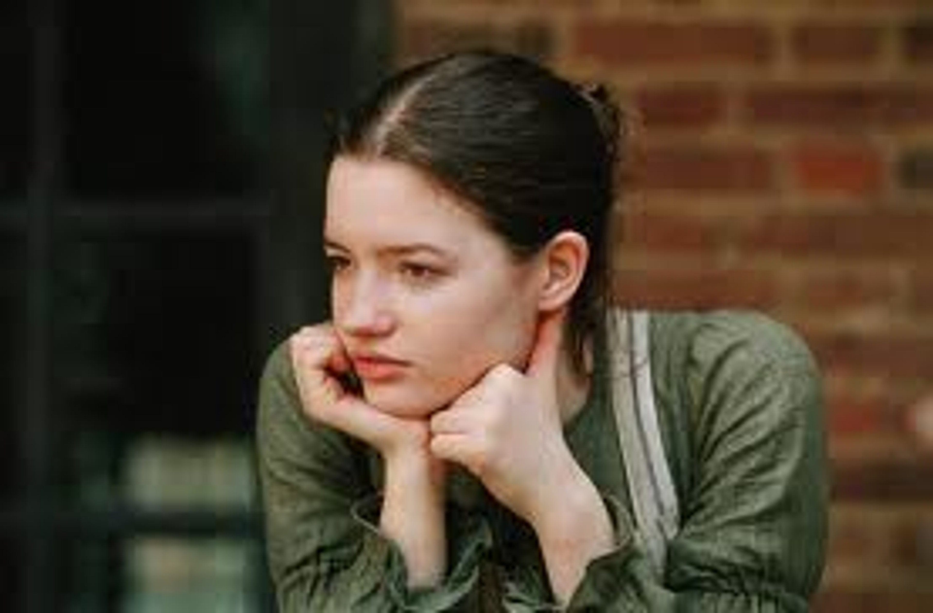 A young woman with dark hair wearing a gray sweater and sitting in front of a brick wall.