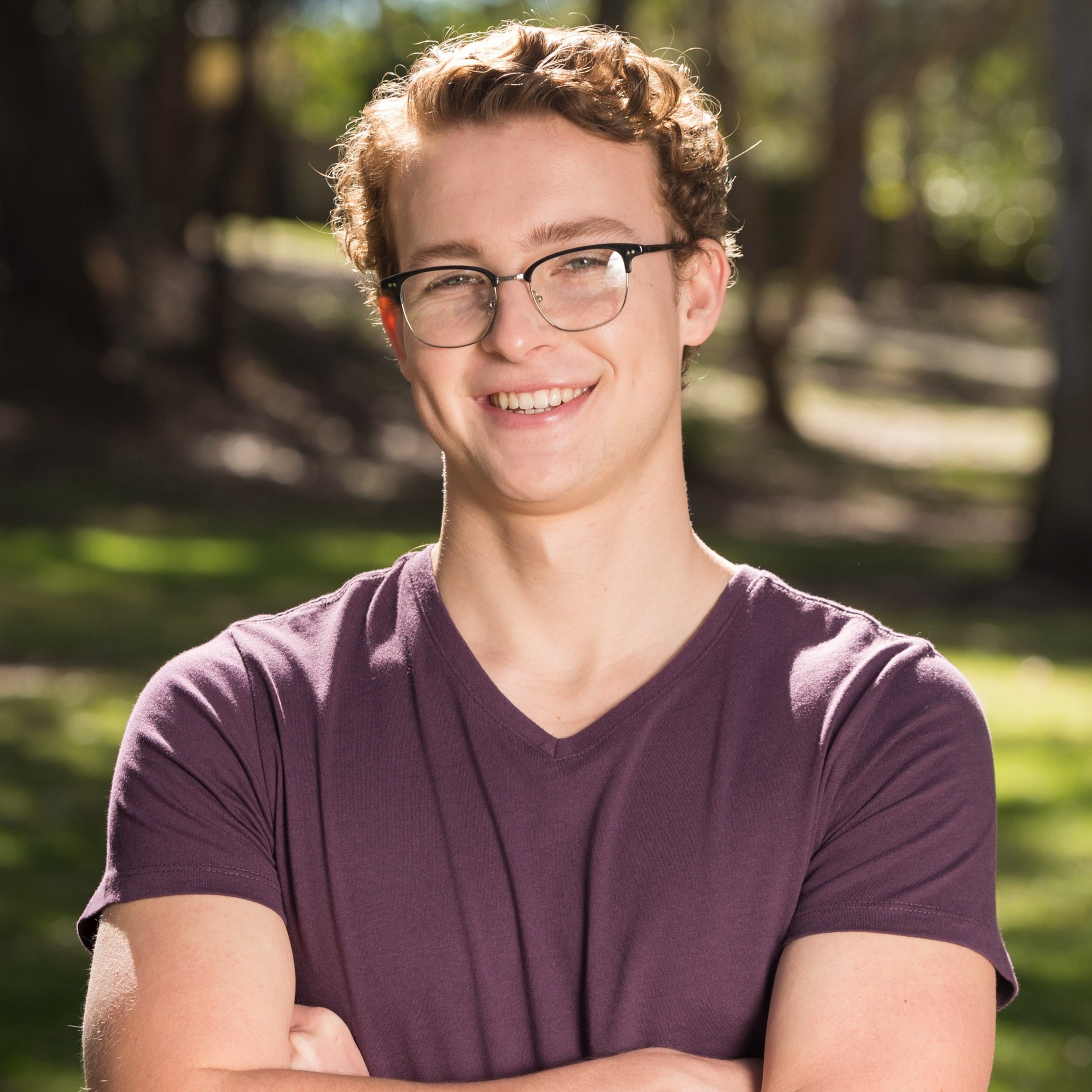 A young man with curly brown hair and glasses, smiling at the camera.