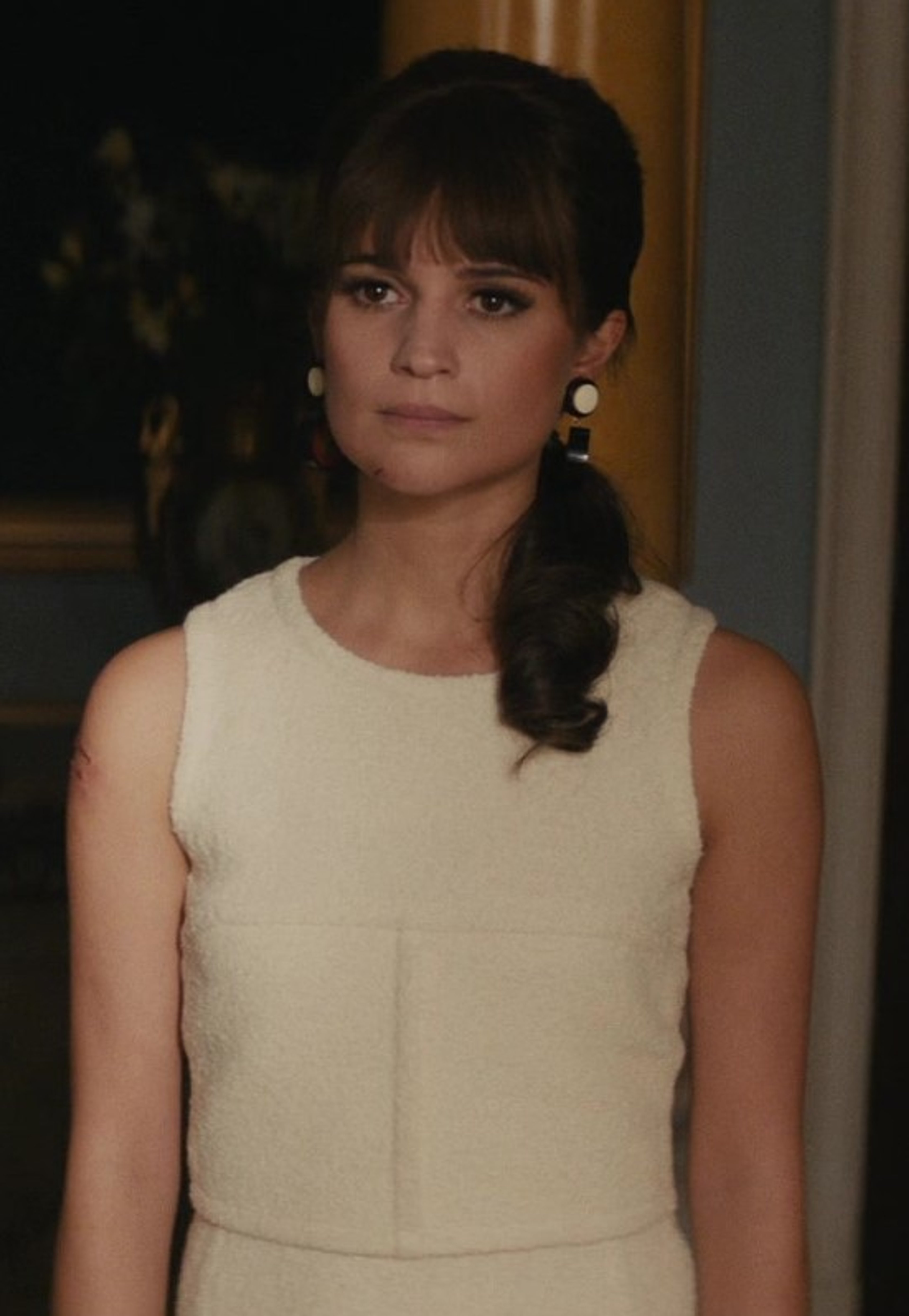 A young woman with dark hair in a braid, wearing a light-colored sleeveless dress, with a serious expression on her face.