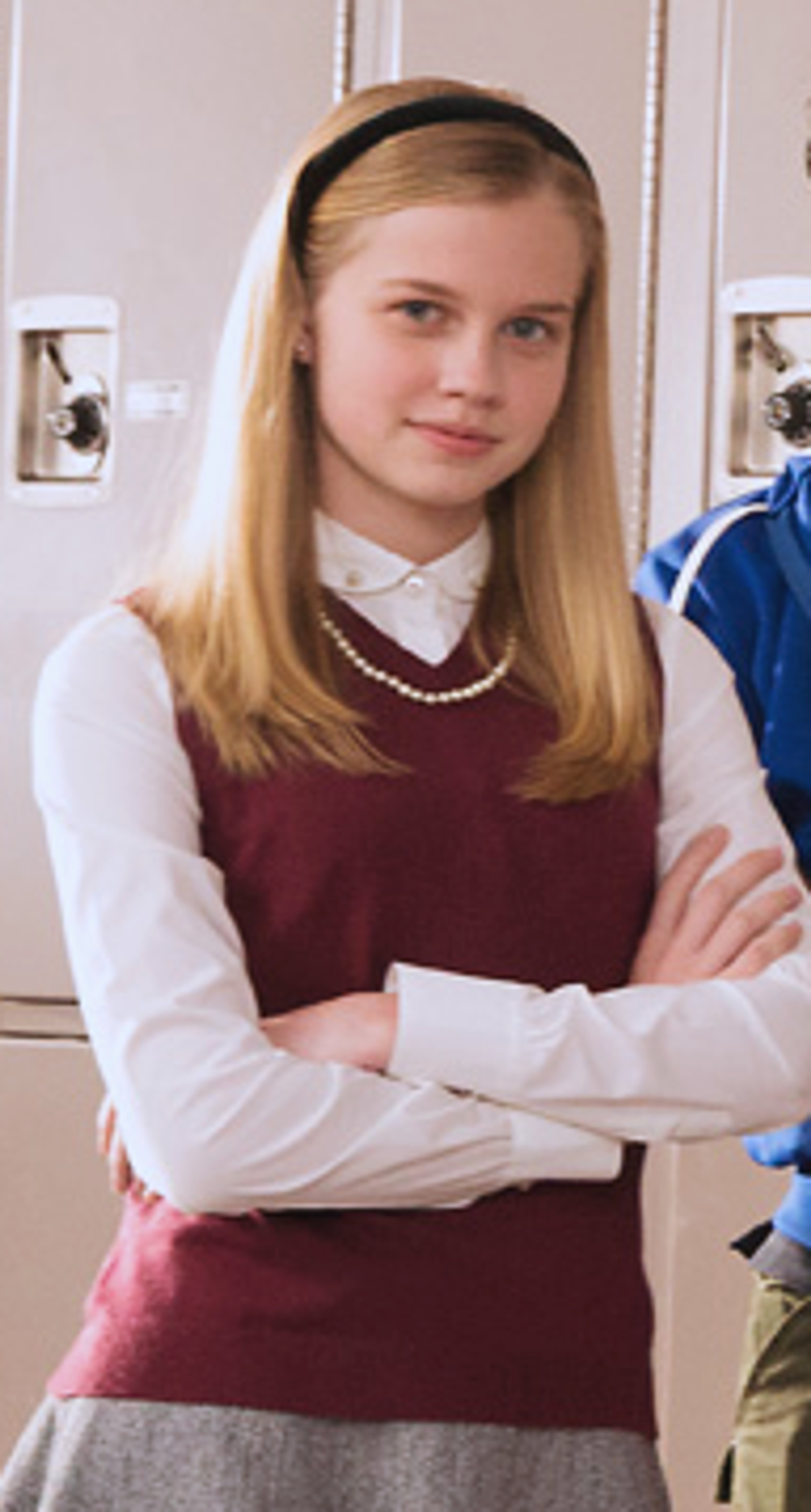 A young woman with blonde hair wearing a burgundy sweater vest and white collared shirt, standing in a school setting with a serious expression.