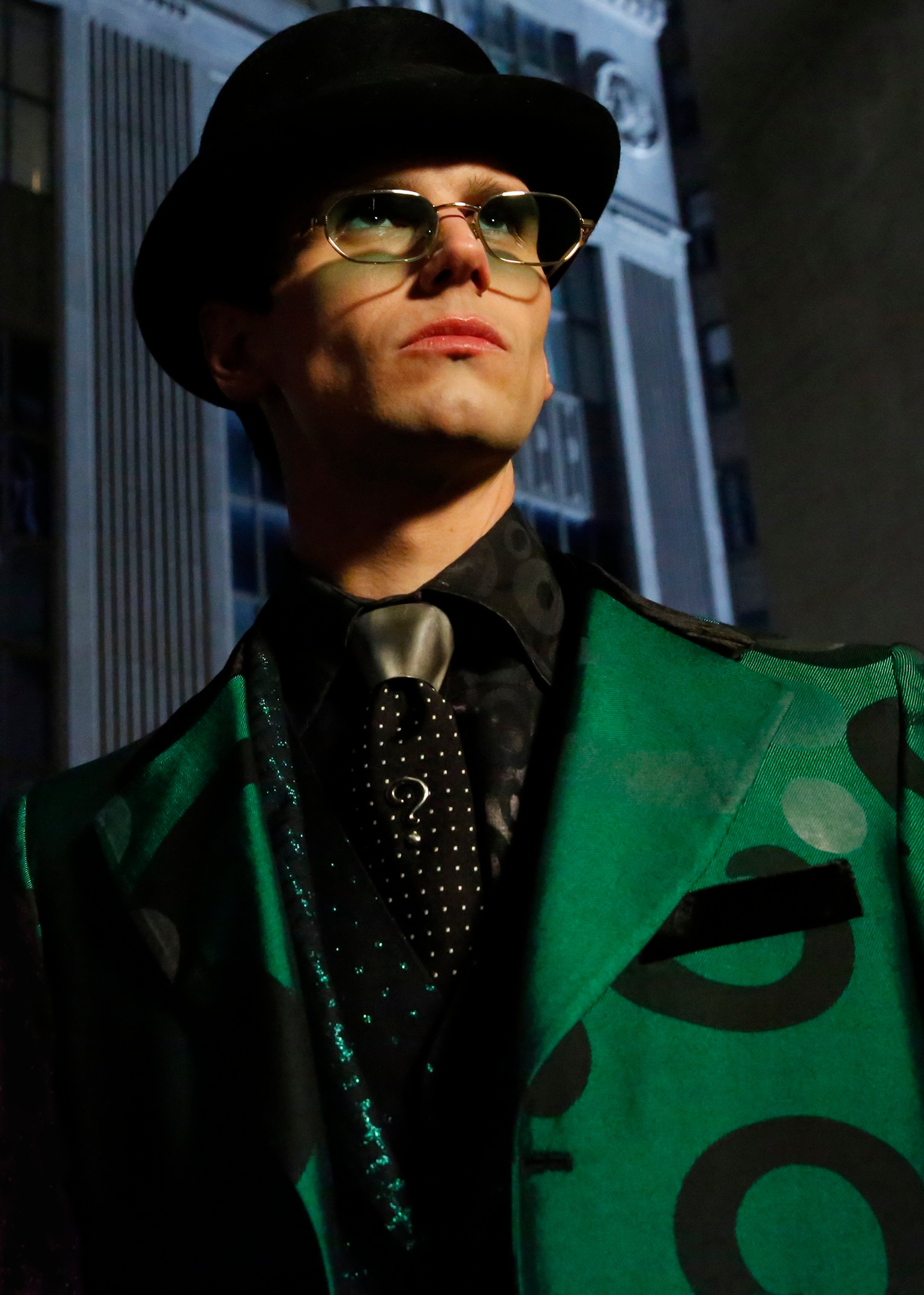 A man in a green and black patterned coat, black hat, and sunglasses standing in front of a building