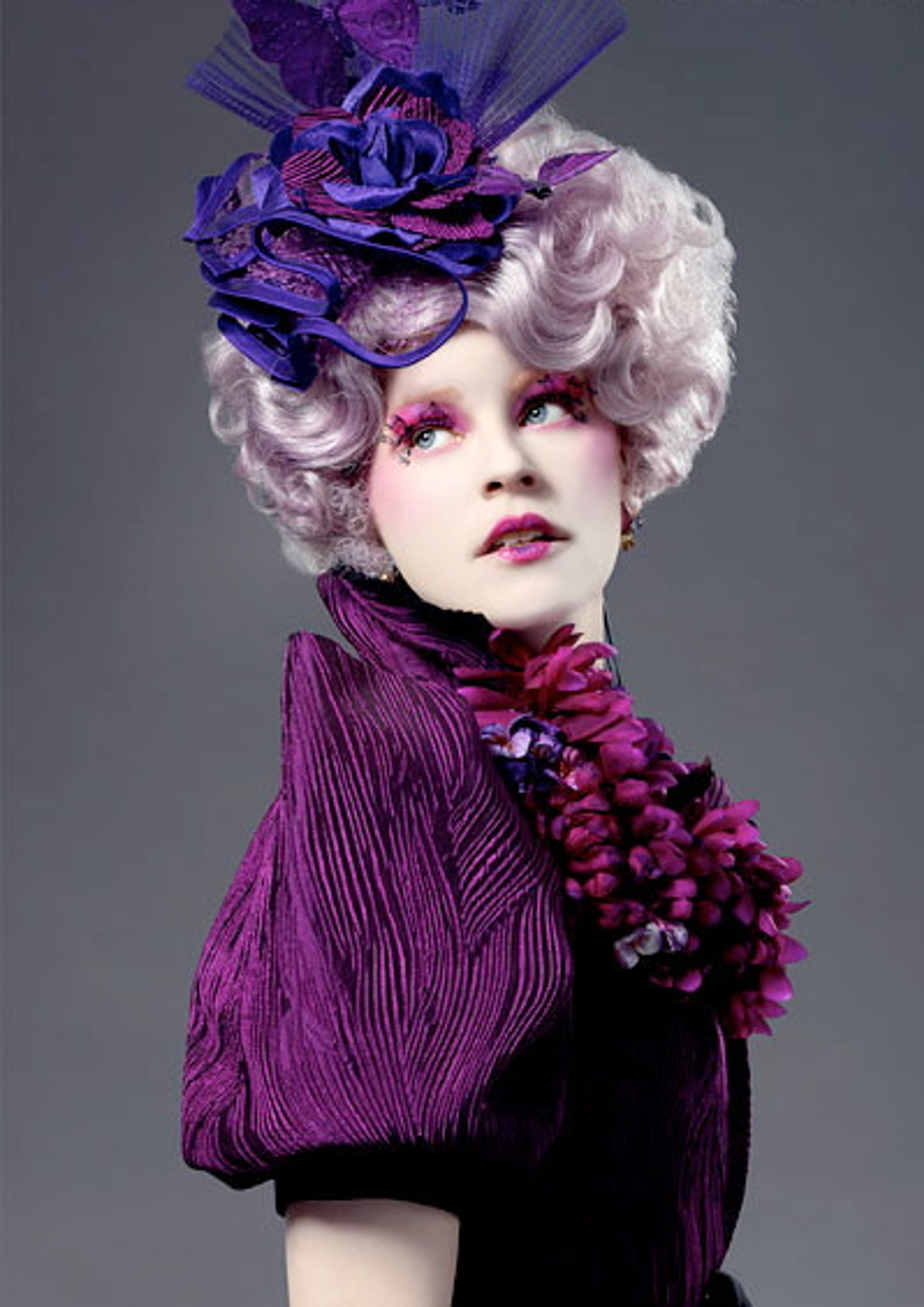 A woman with an elaborate, colorful makeup and hairstyle wearing a large, ornate purple hat with feathers and flowers.