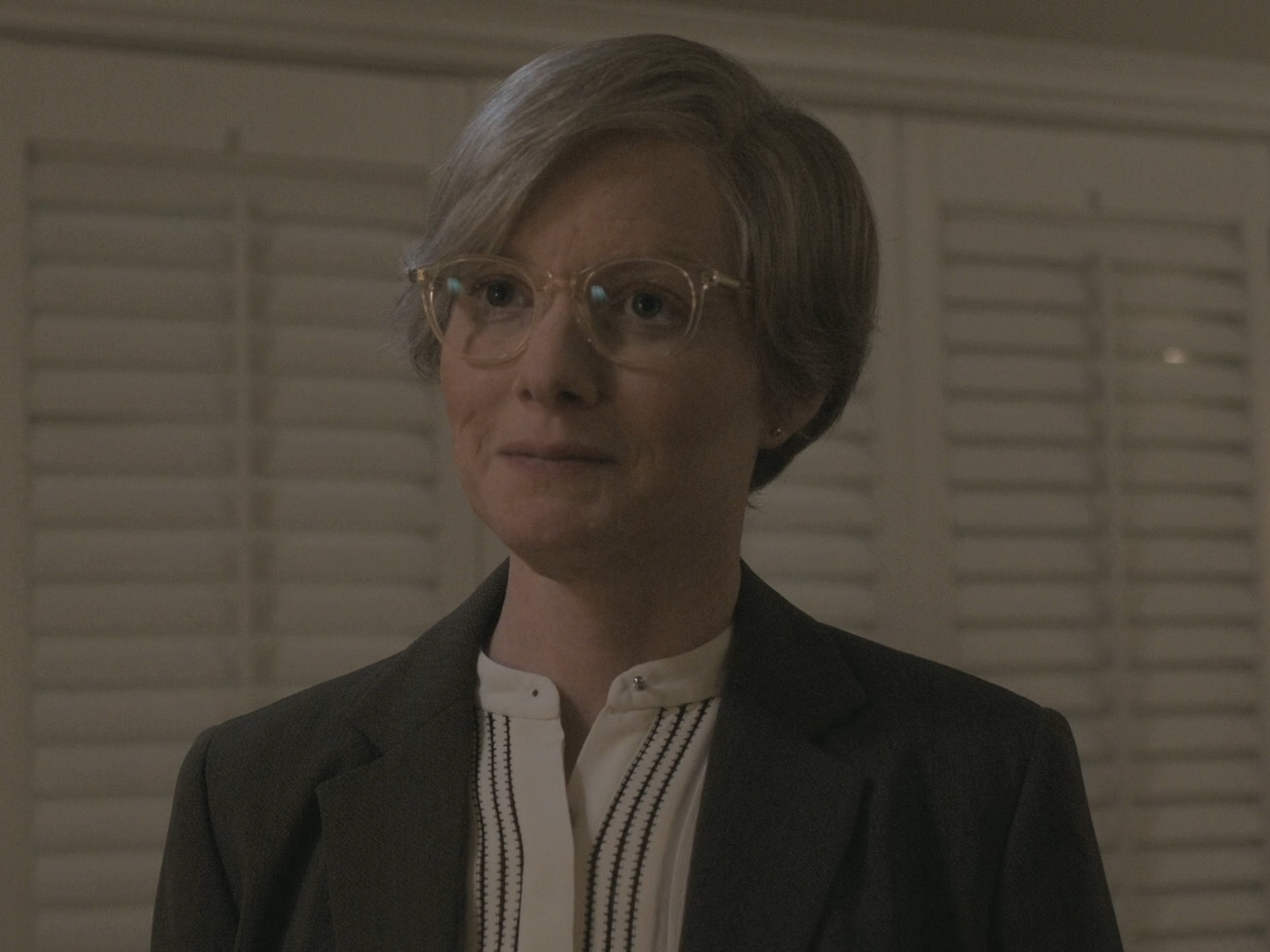 A middle-aged woman with short gray hair and glasses, wearing a black blazer and white blouse, with a serious expression.