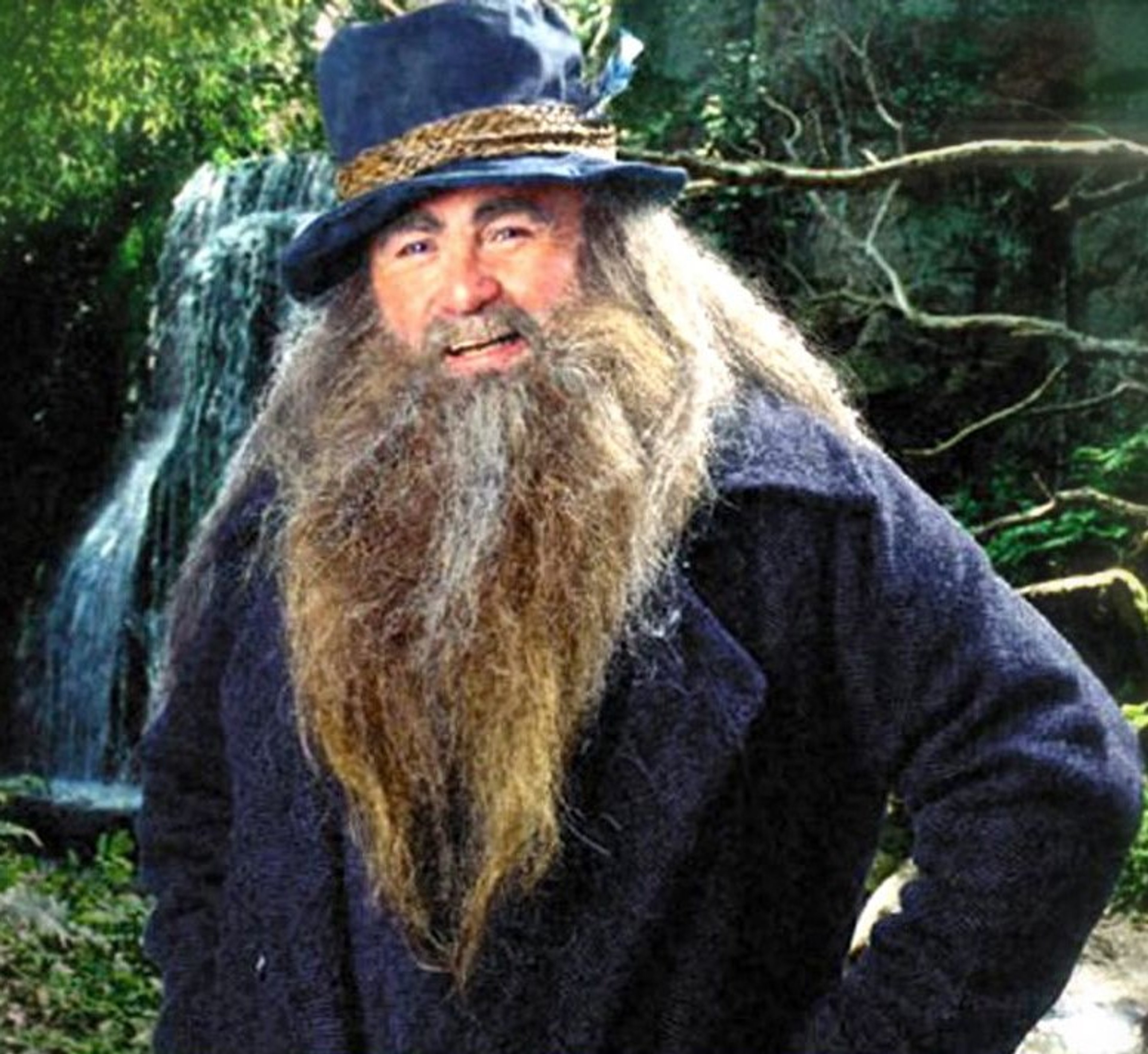 An elderly man with a long white beard and hair, wearing a wide-brimmed hat and a dark cloak, standing in a lush, forested environment with a waterfall in the background.