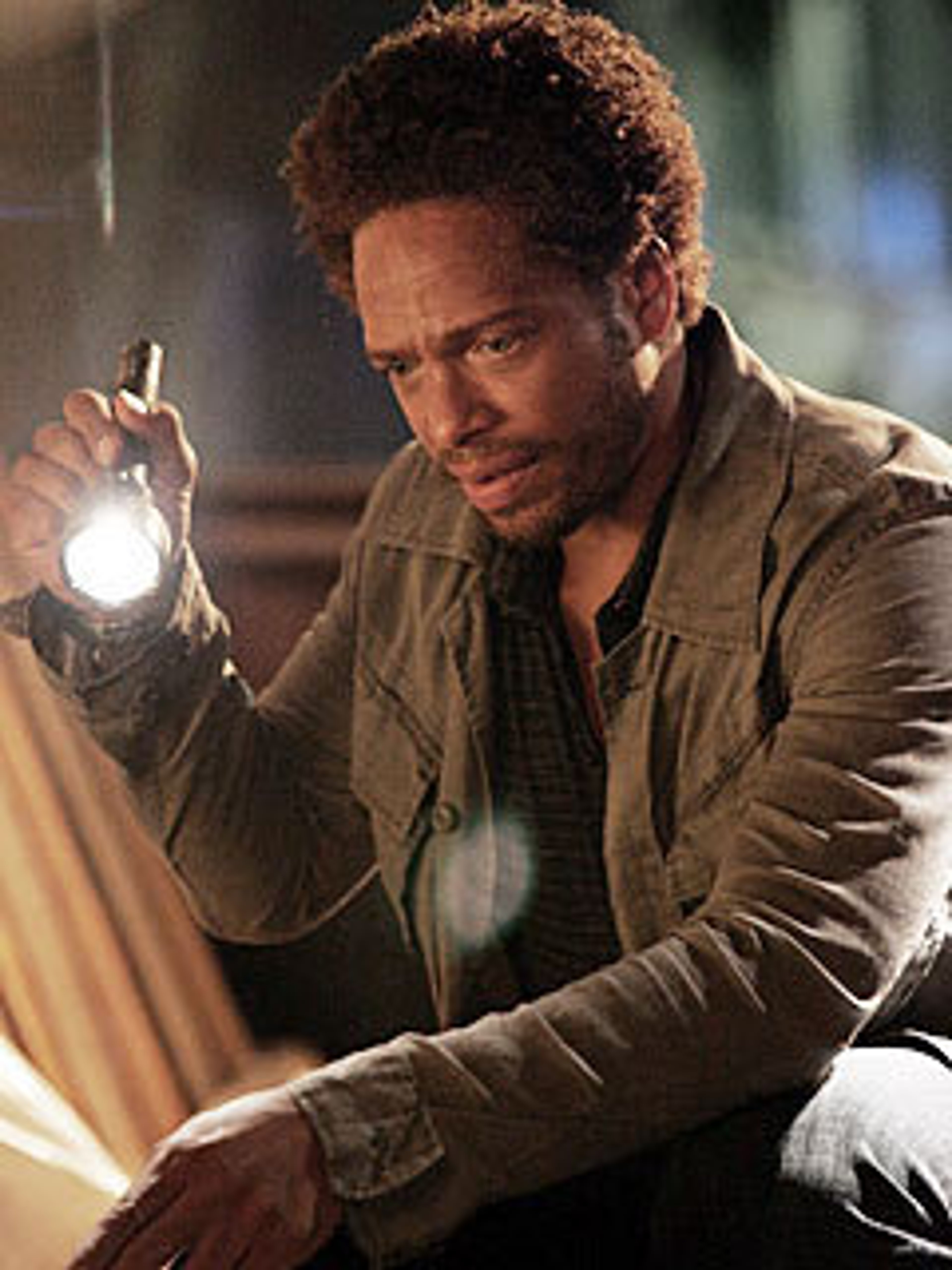 A man with curly hair and a beard holding a flashlight in a dimly lit indoor setting