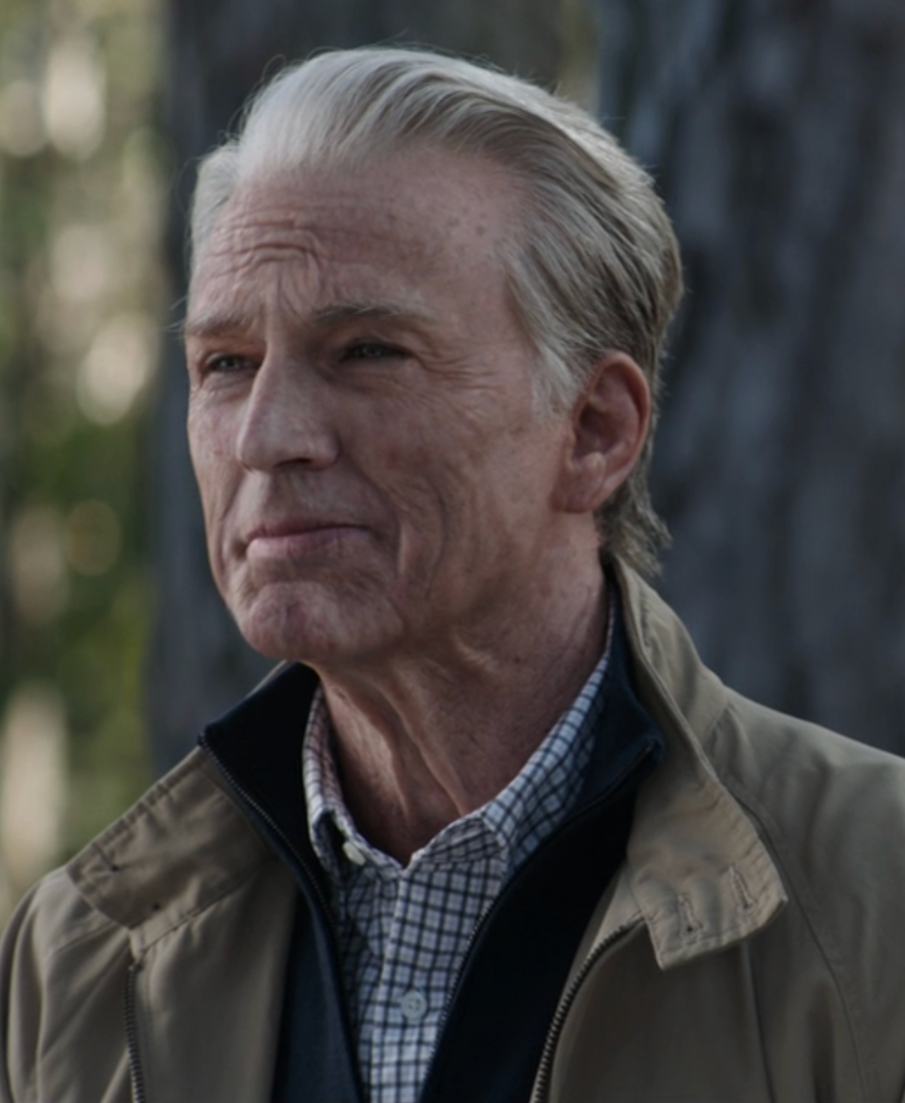 An older man with gray hair and a serious expression, wearing a tan jacket and plaid shirt.