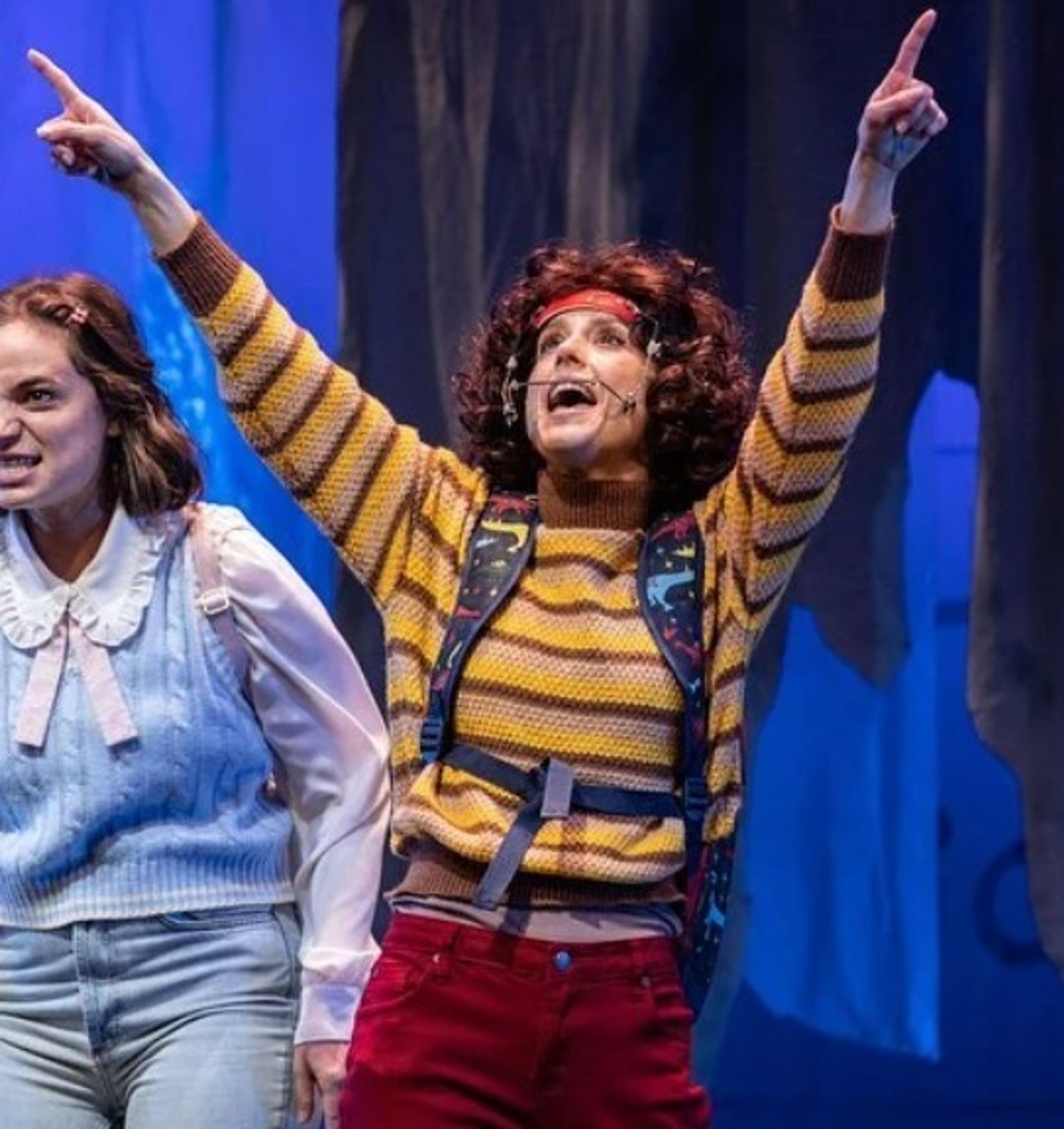 A young woman with curly brown hair in a celebratory pose, wearing a yellow and black striped sweater and red pants.