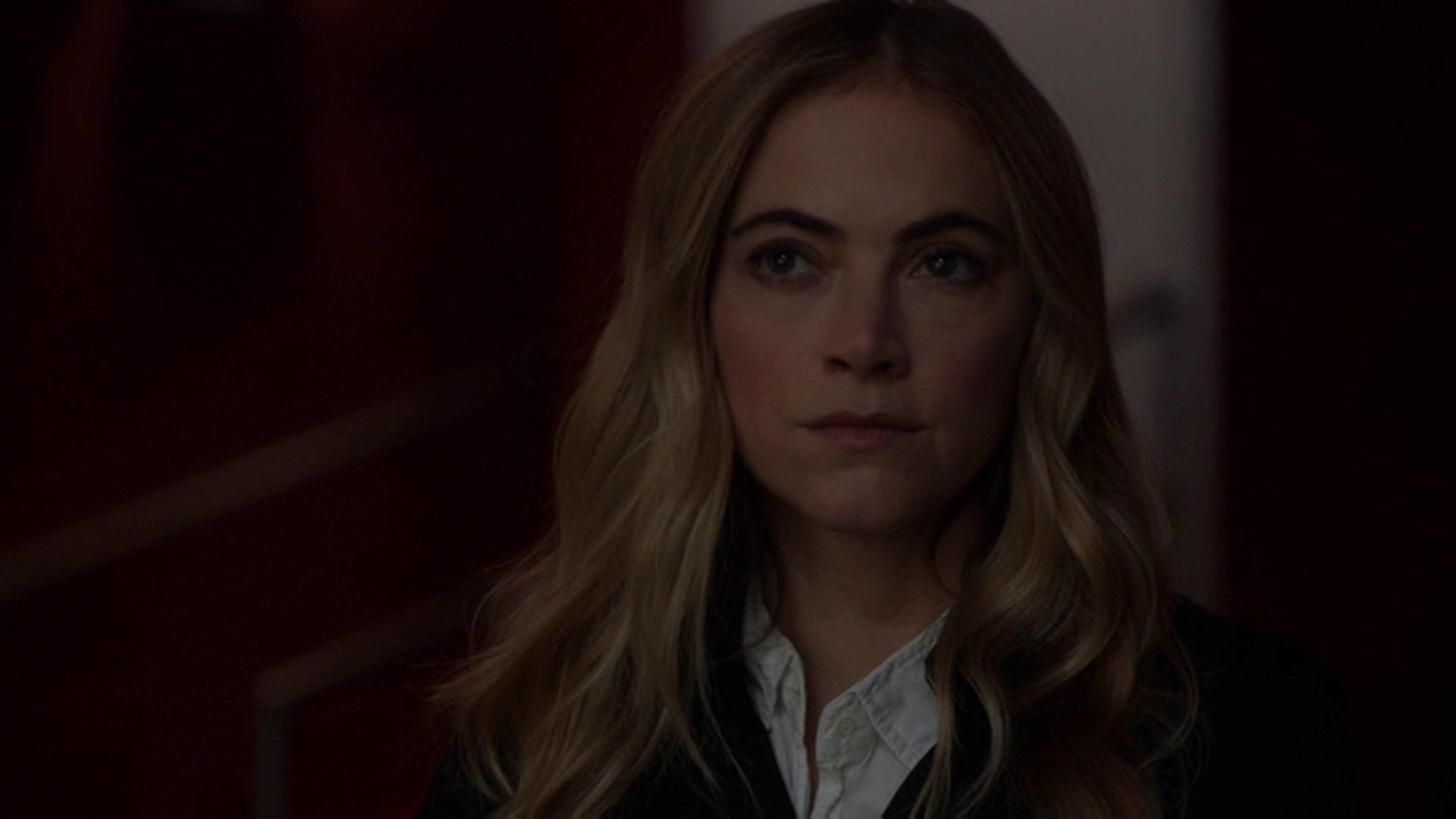 A woman with long blonde hair wearing a white collared shirt in a dimly lit setting.