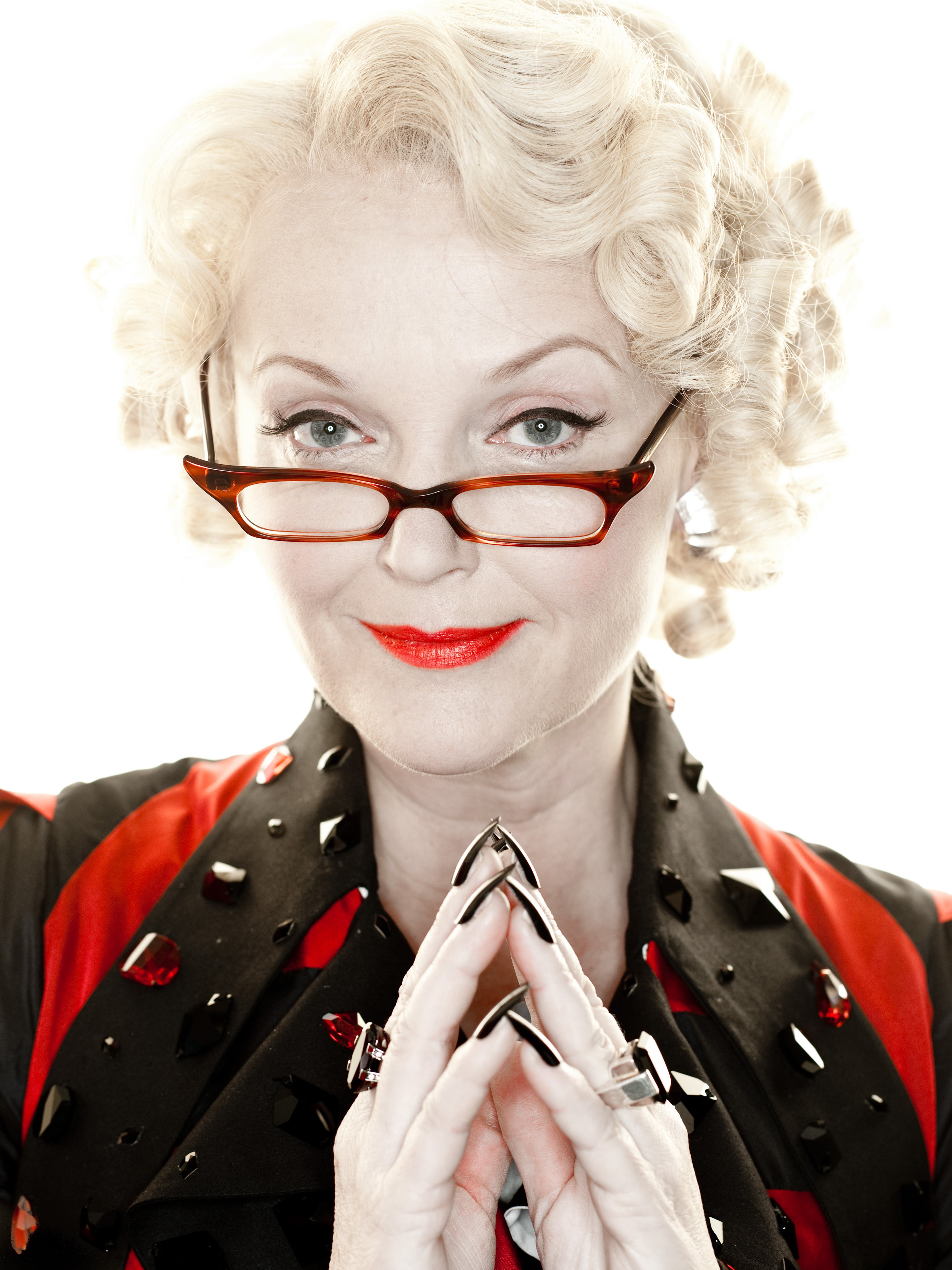 A woman with blonde curly hair wearing glasses and a polka dot outfit making a praying gesture