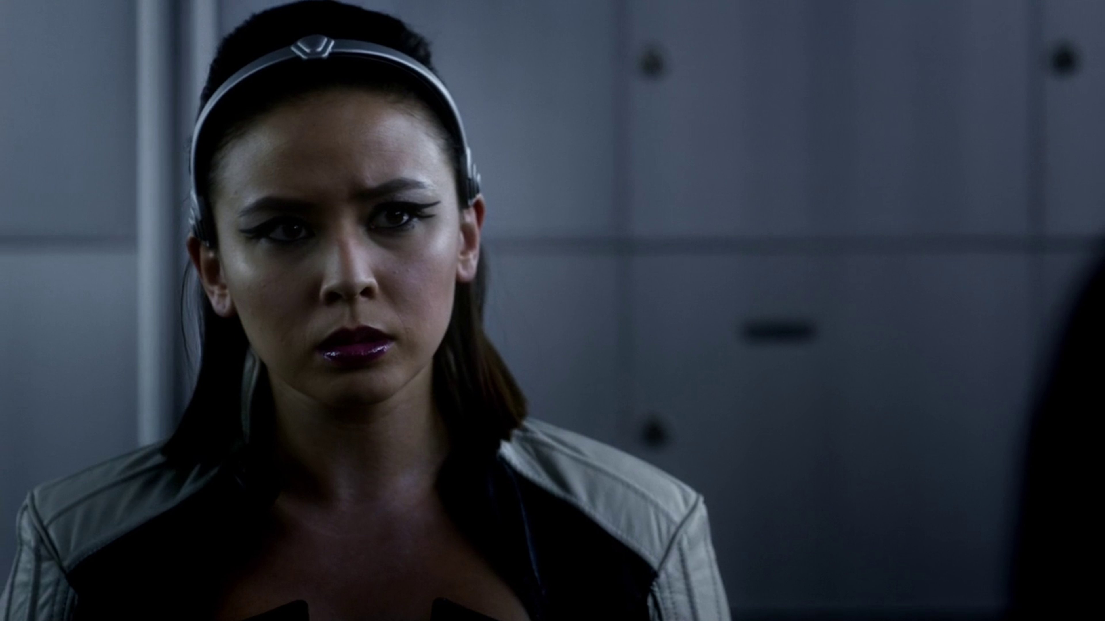 A woman with dark hair wearing a black outfit, looking serious and intense.