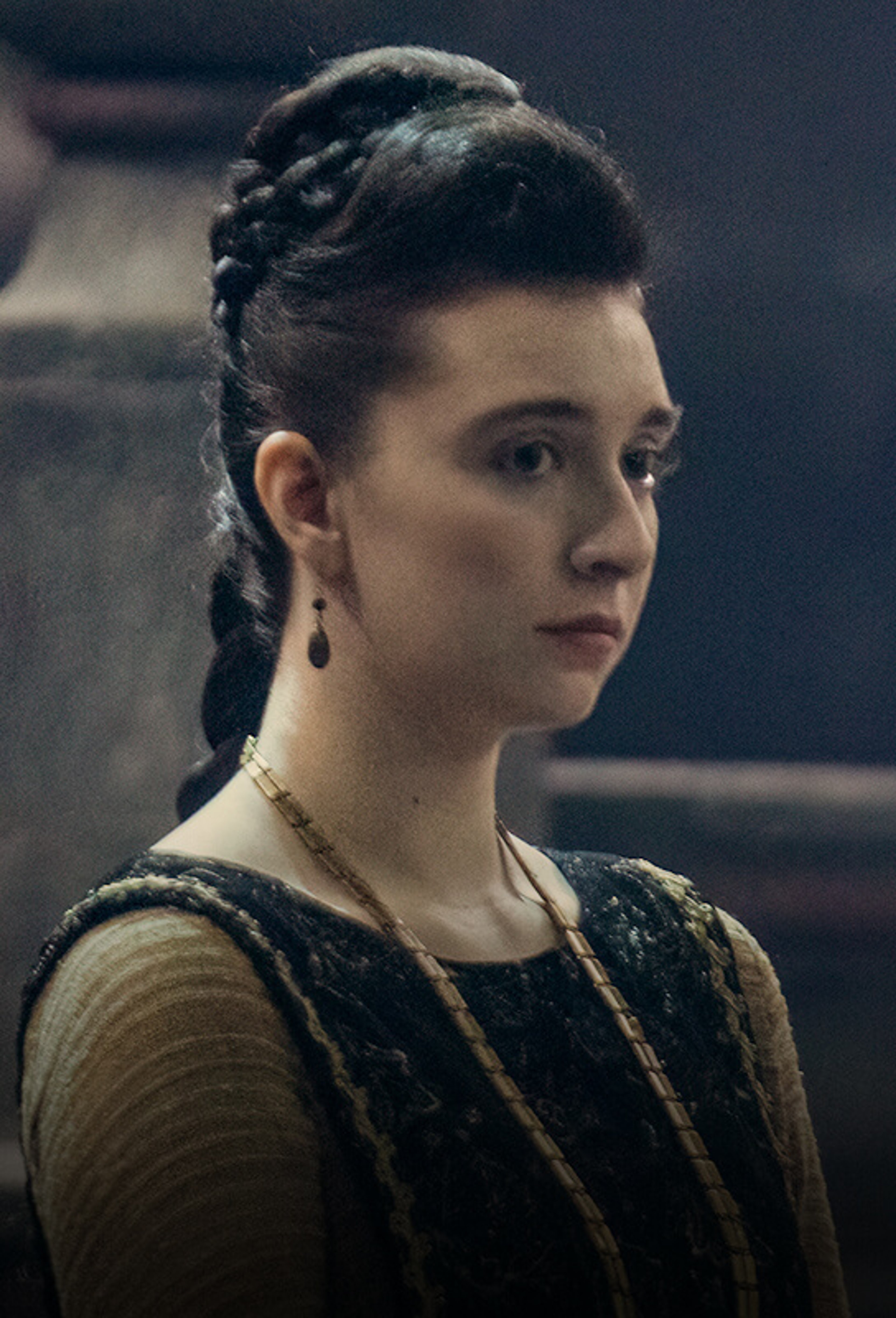 A young woman with dark hair and a serious expression, wearing a dark dress with gold accents.