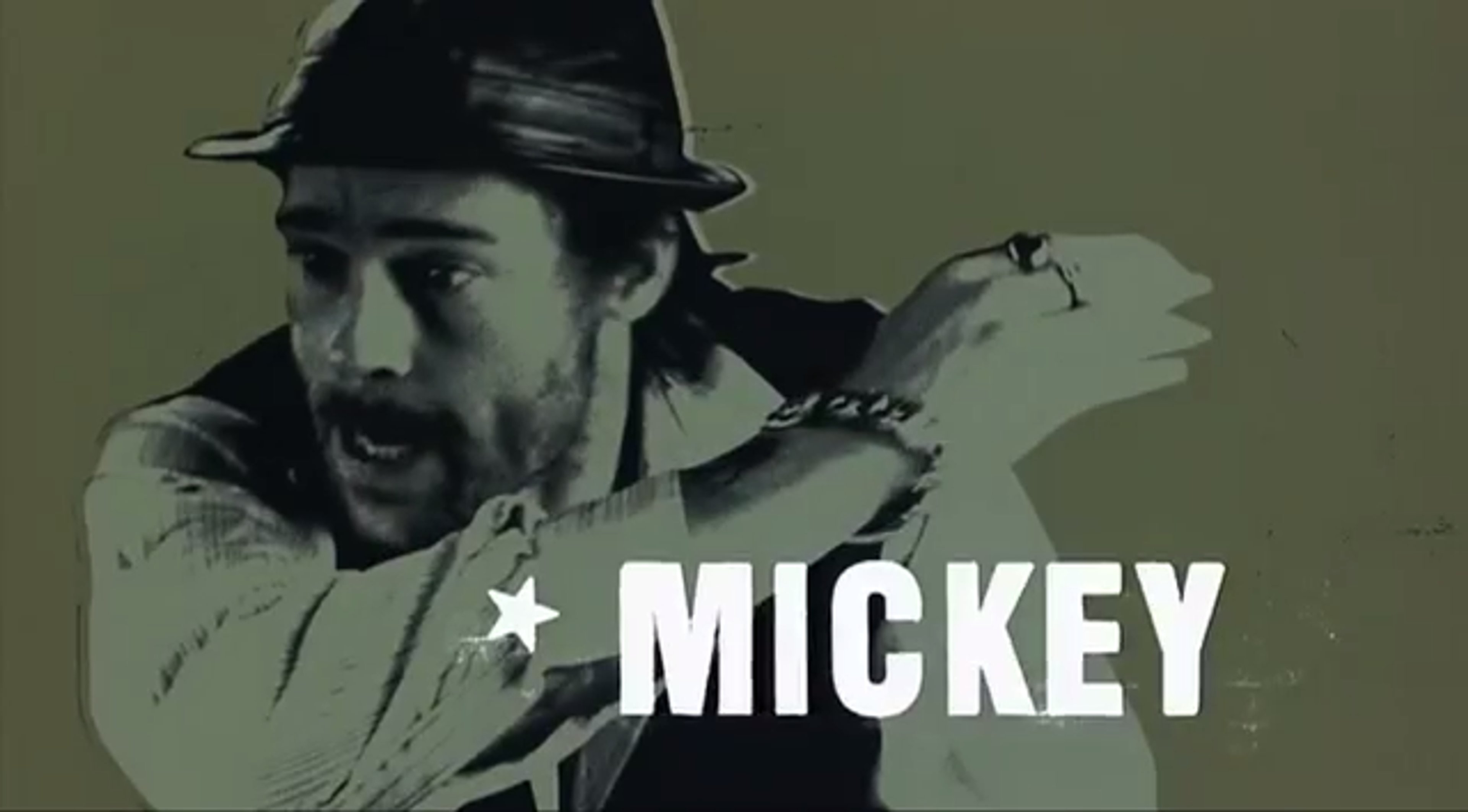 A middle-aged man with a rugged appearance wearing a black hat and a shirt with the word 'MICKEY' printed on it.