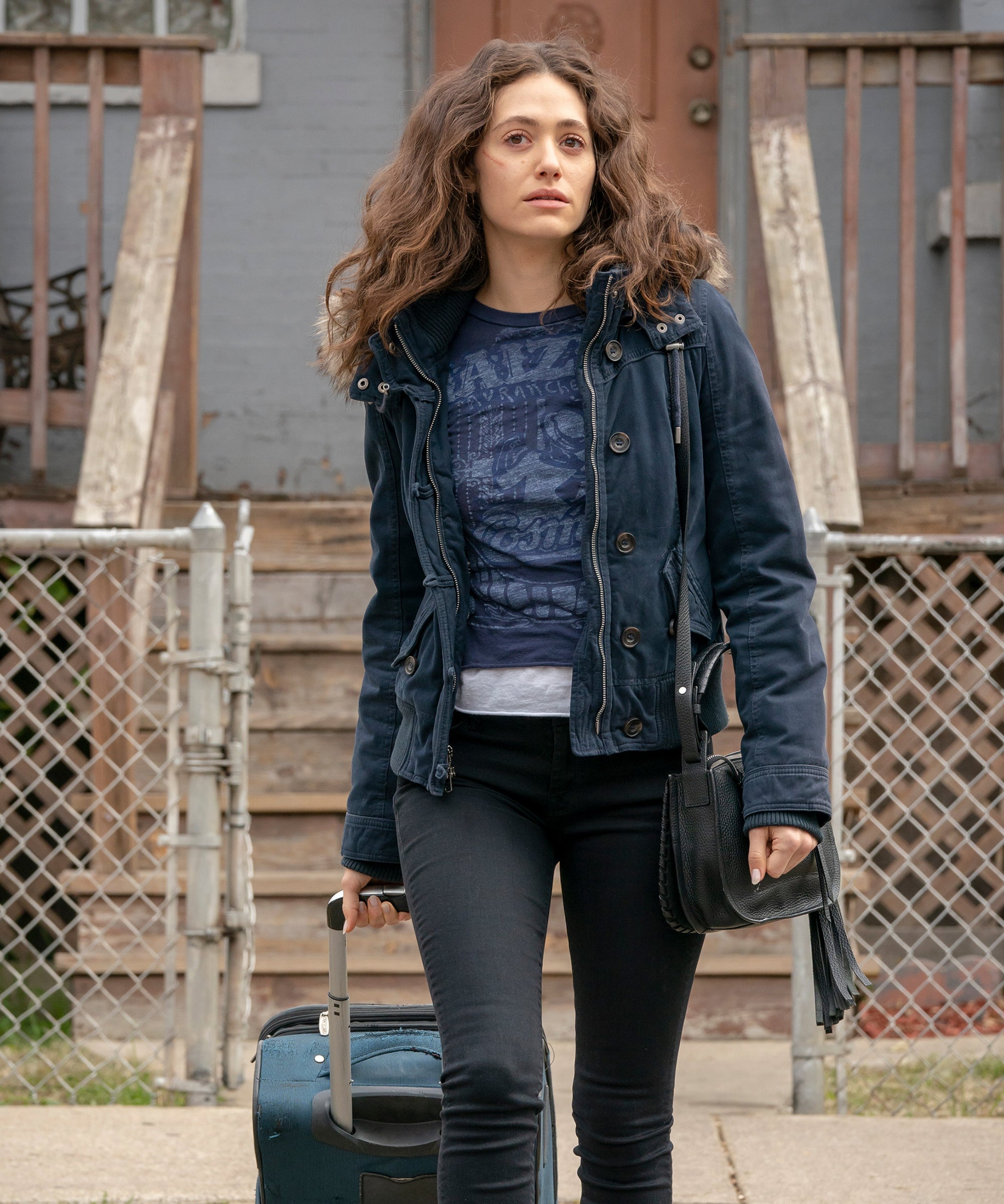 A young woman with curly brown hair wearing a black jacket and jeans, standing in front of a fence or gate.