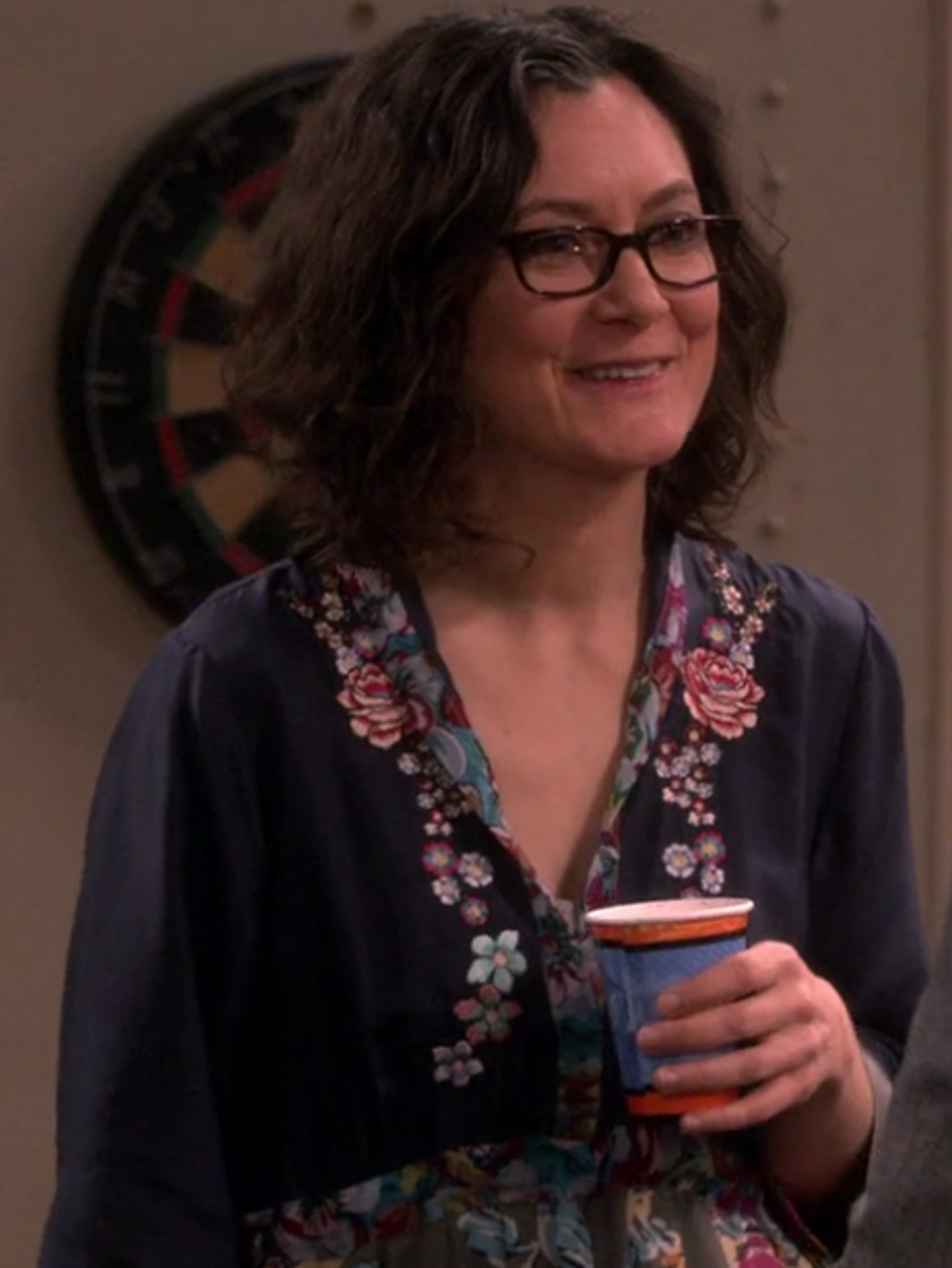 A woman with curly brown hair wearing glasses and a floral patterned shirt, smiling and holding a mug or cup.