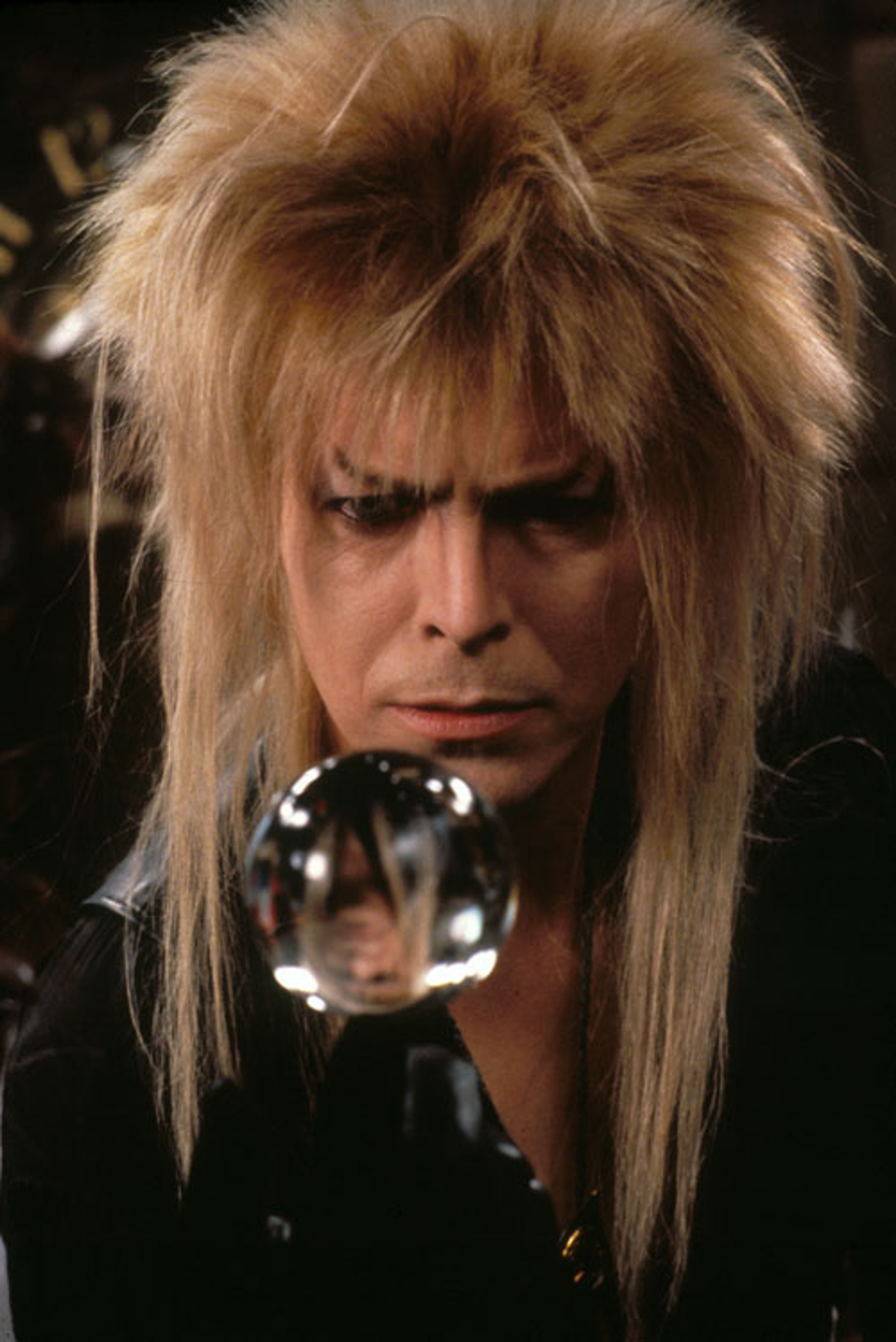 A man with wild blonde hair and heavy eye makeup holding a crystal ball, likely the Goblin King Jareth from the film Labyrinth.