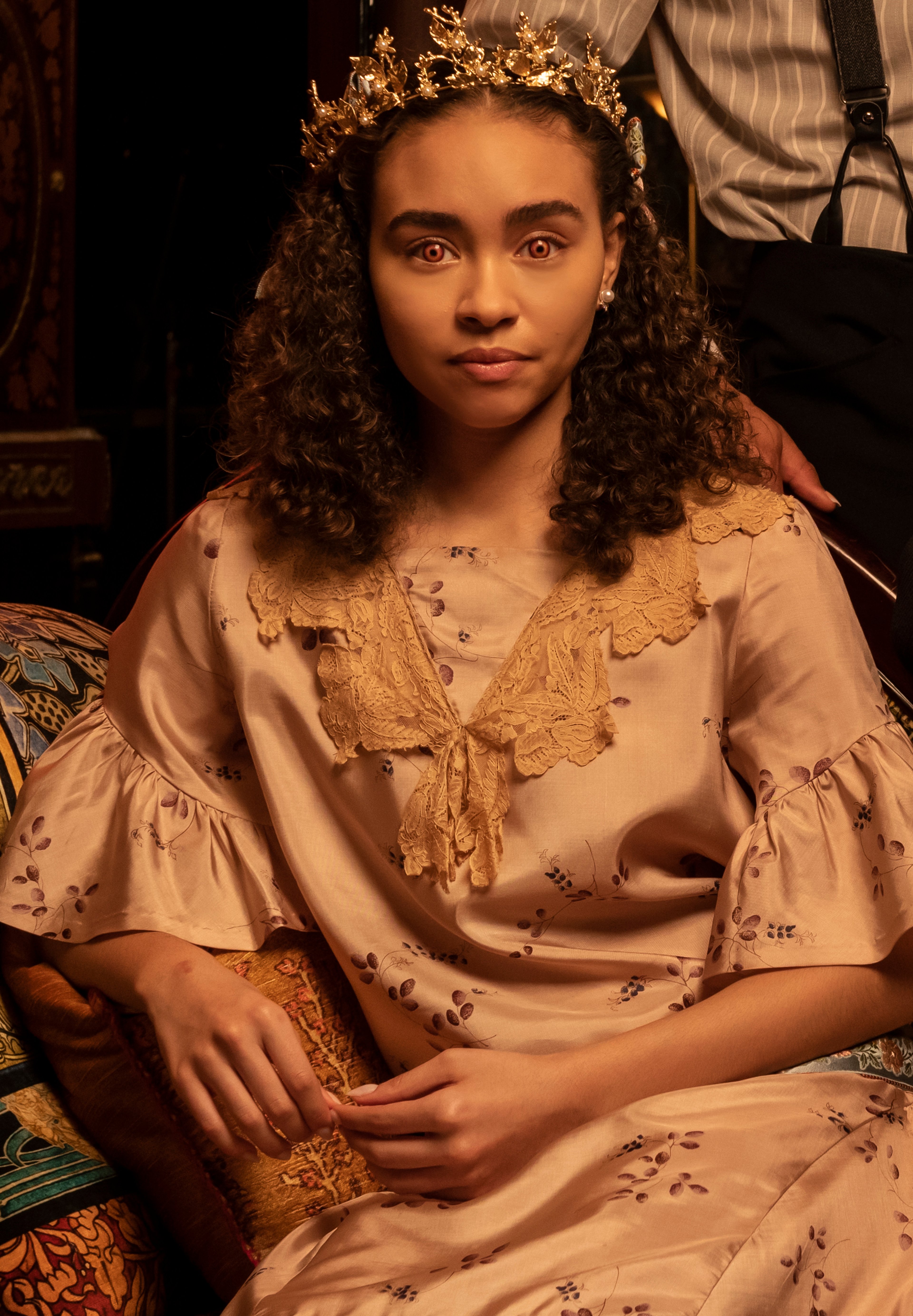 A young woman with curly dark hair wearing a beige dress and a floral crown
